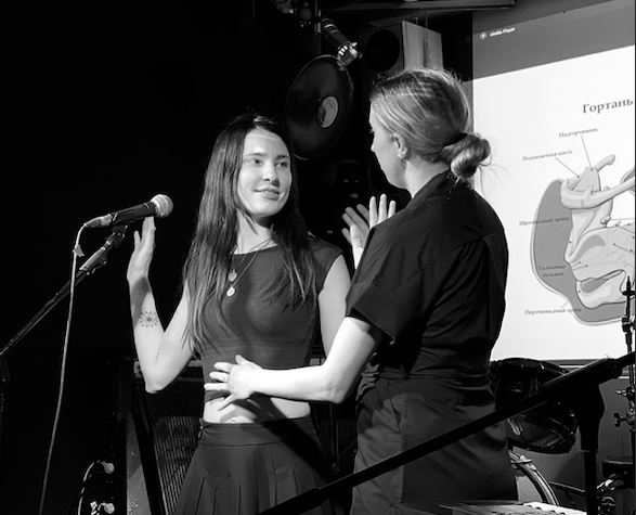 Two women in conversation during a presentation or performance, with a microphone on a stand and a projected diagram of the human digestive system in the background.