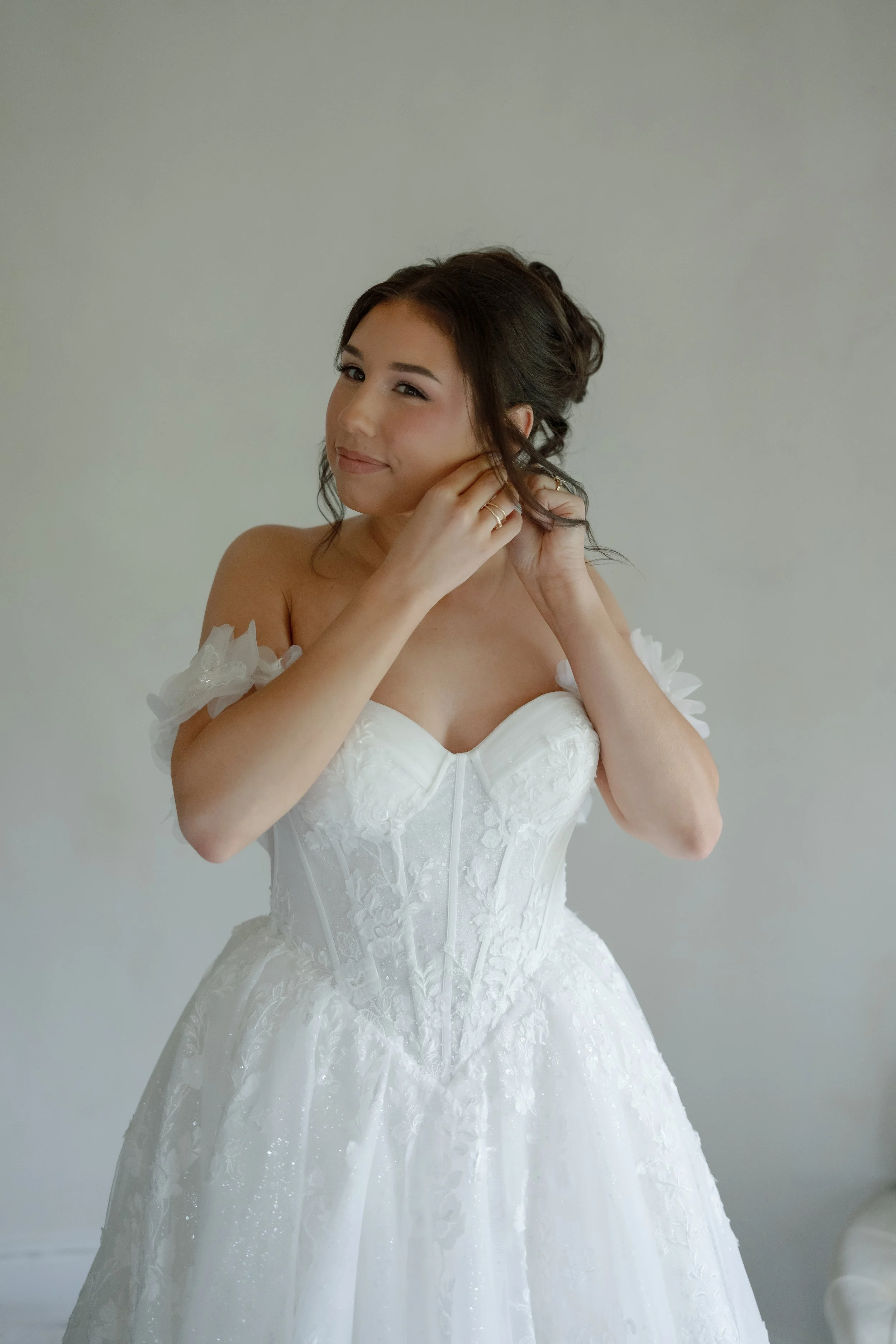 Bride in a white lace wedding dress adjusting her hair against a light gray background