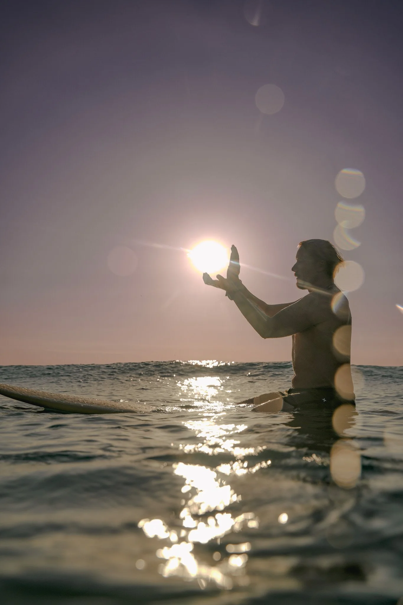 Surf photographer capturing a surfer in the Atlantic Ocean during the August 2026 partial solar eclipse in Cornwall, UK. The setting sun is almost completely obscured by the Moon as the surfer sits in the water, creating dramatic silhouettes, reflect