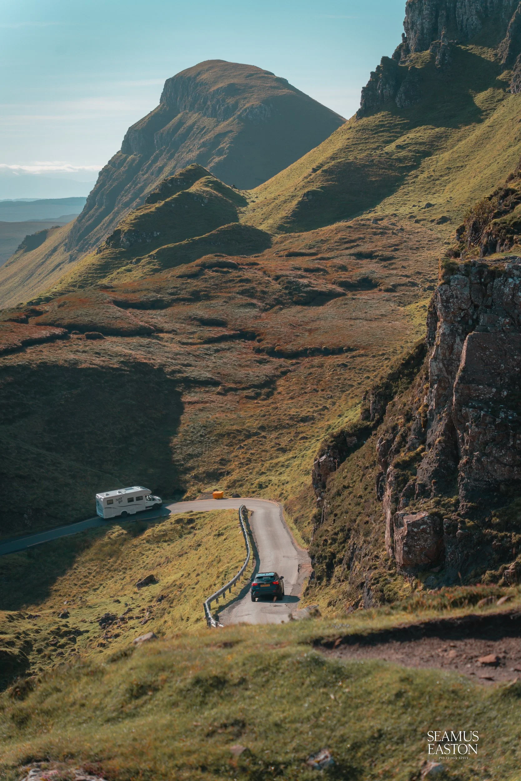 A winding mountain road with a black car, a white camper van, and an orange box on the road, surrounded by green hills and rugged cliffs under a clear blue sky.