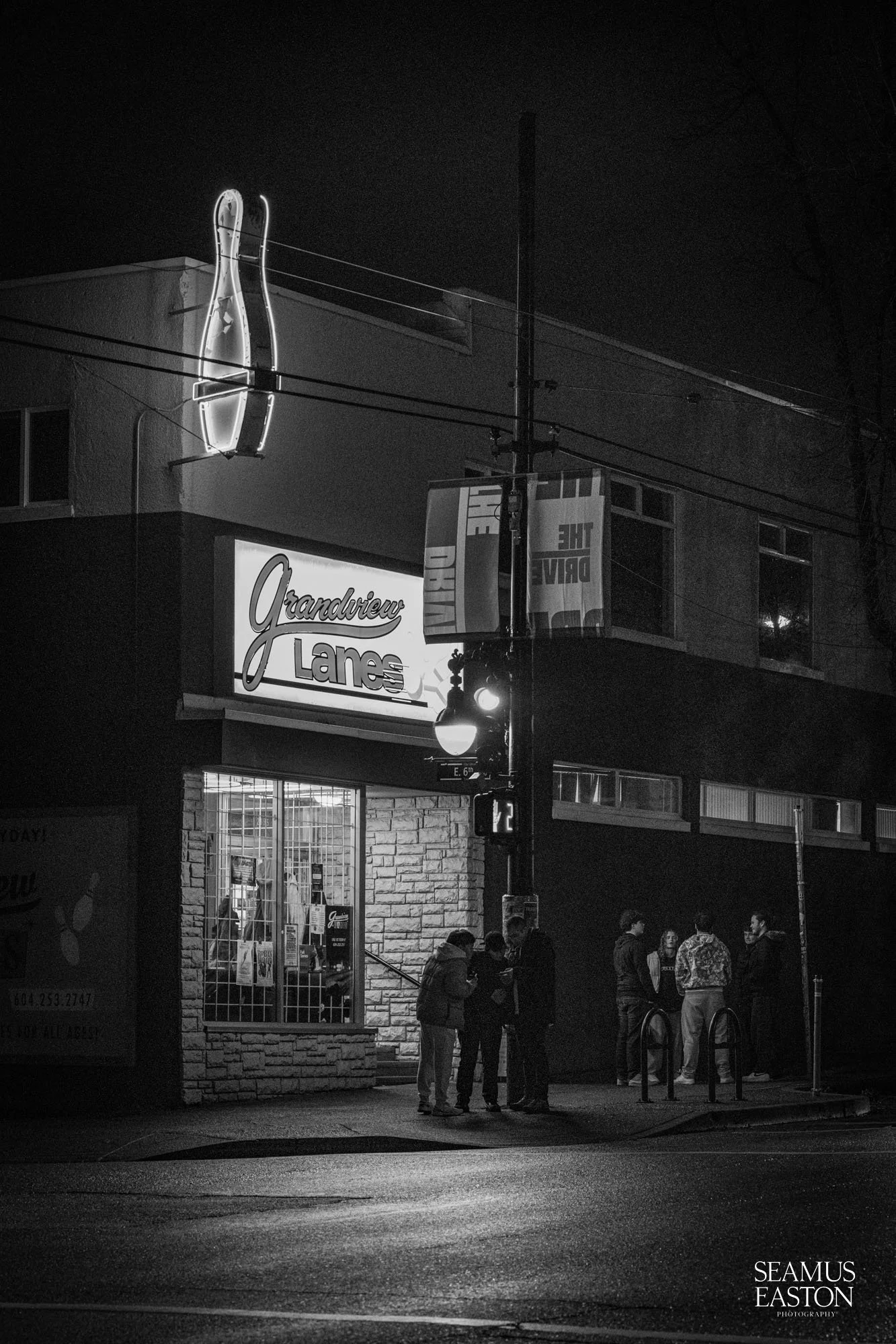 Nighttime scene with a group of people standing outside Grandview Lanes bowling alley, illuminated sign with a bowling pin and ball, brick facade, and streetlights.