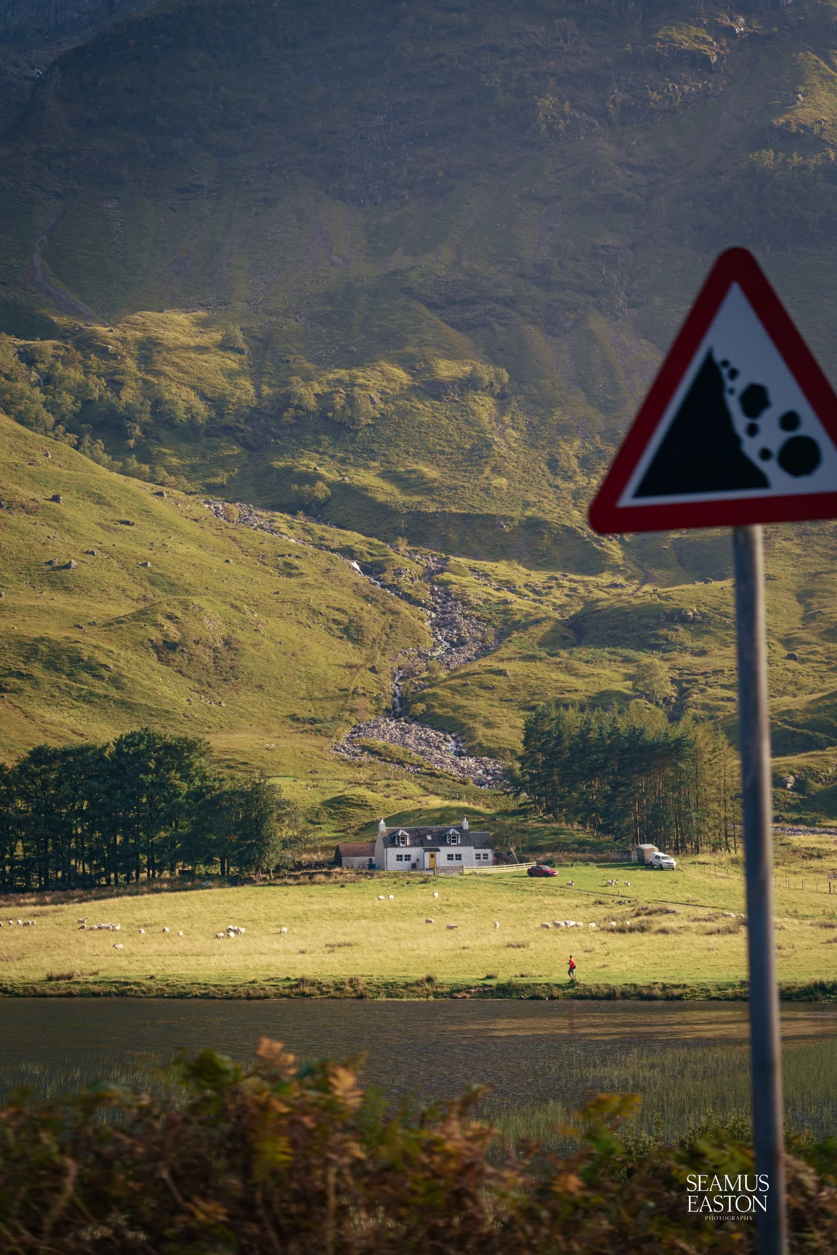Countryside with green hills, trees, a house, a person walking, and a sign warning of falling rocks.