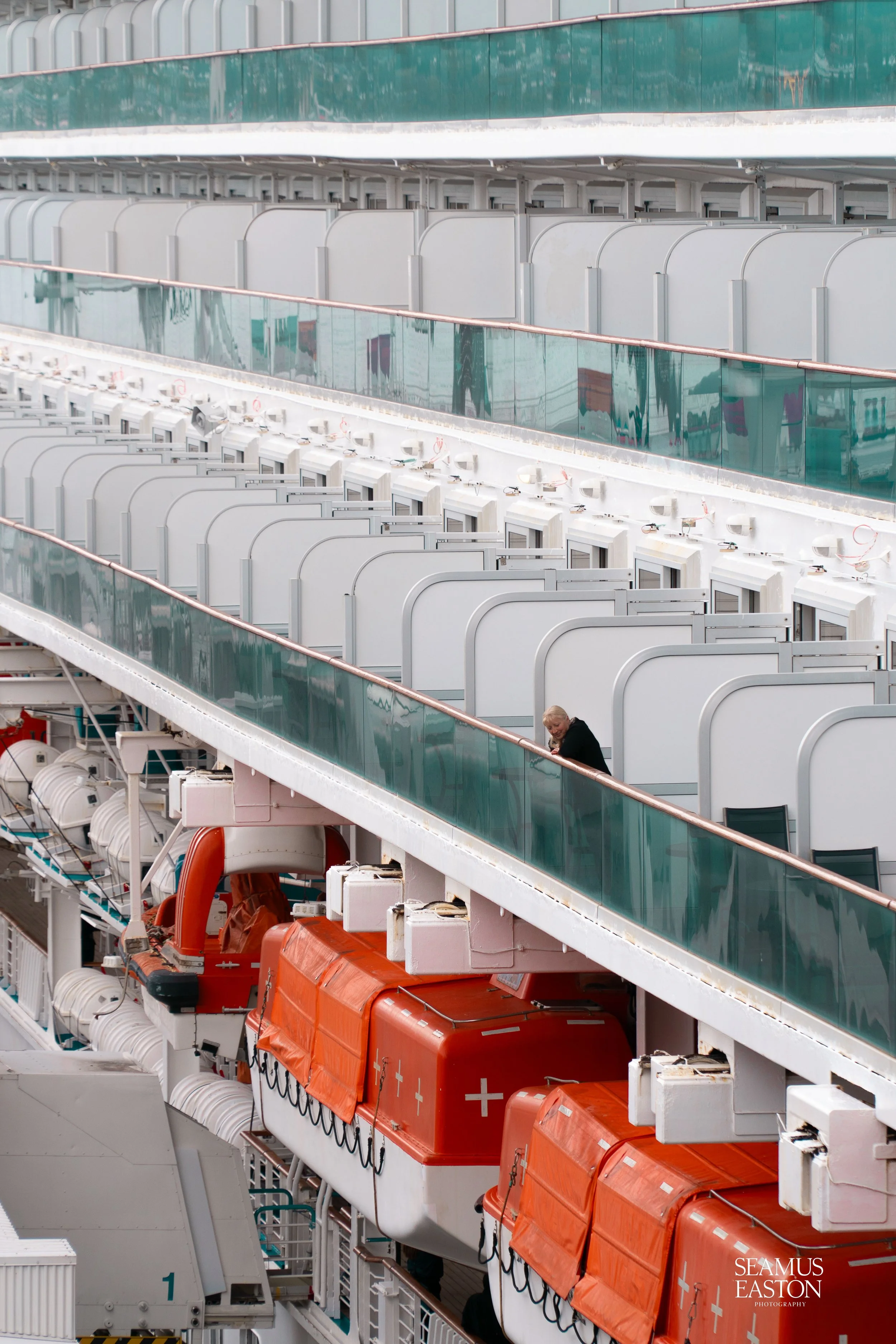 An interior view of the upper decks of a cruise ship, showing rows of hallway doors and deck chairs, with emergency life rafts and equipment in the foreground.