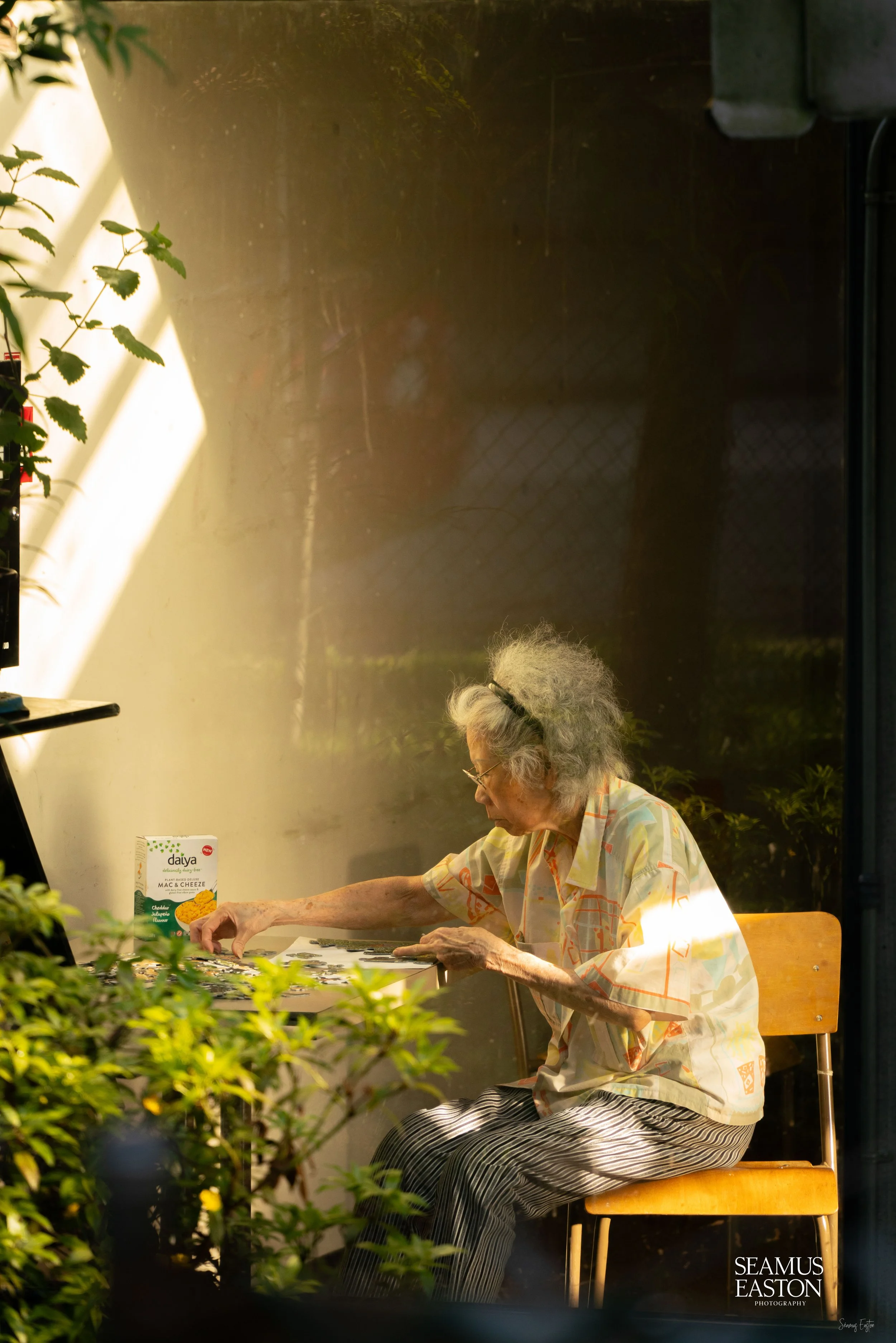 An elderly woman with curly gray hair, wearing glasses, a pastel patterned shirt, and striped pants, is seated outdoors at a small table, playing a puzzle. The scene is softly lit with warm sunlight, surrounded by green plants and garden elements.
