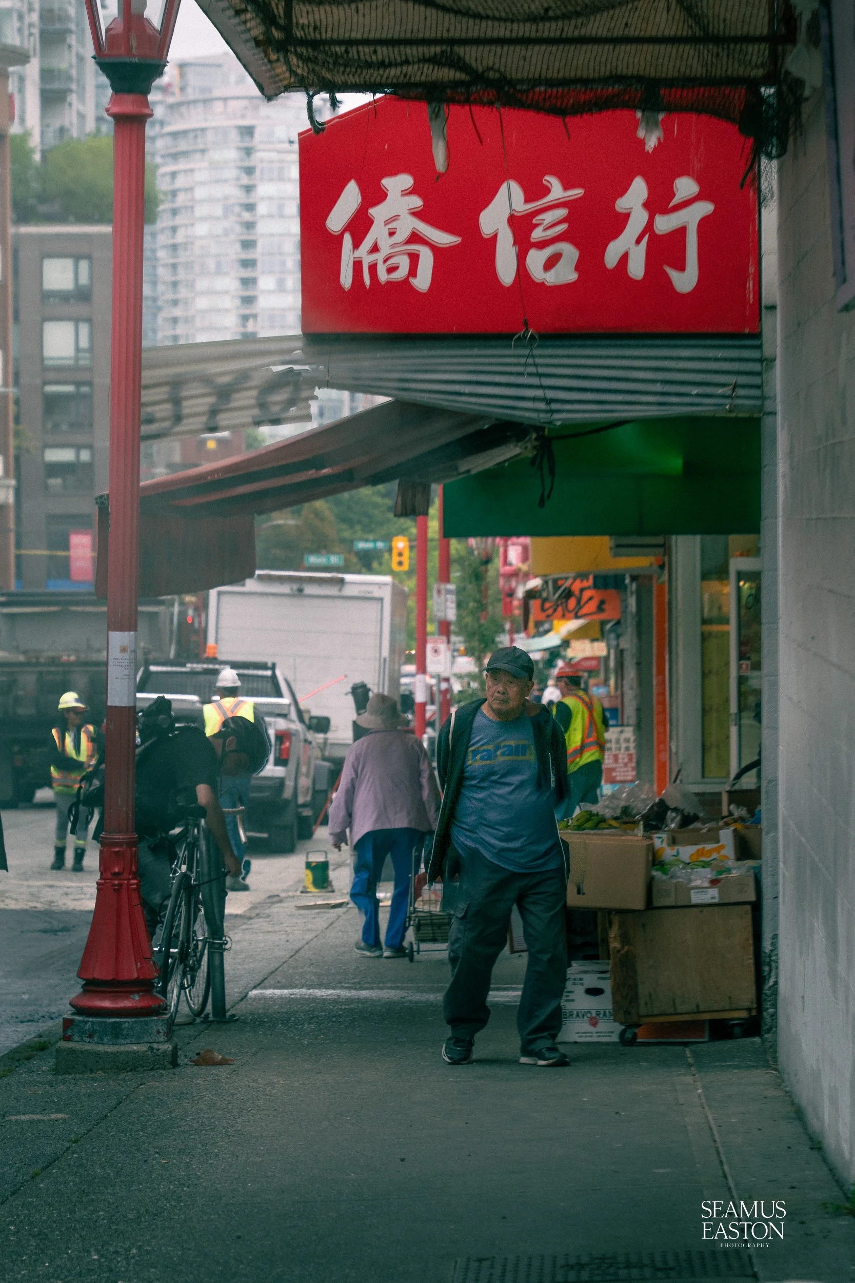 A city street scene with pedestrians, some wearing hats and backpacks, a man walking with hands in pockets, and storefronts with signs in Chinese characters. Buildings and traffic are visible in the background.