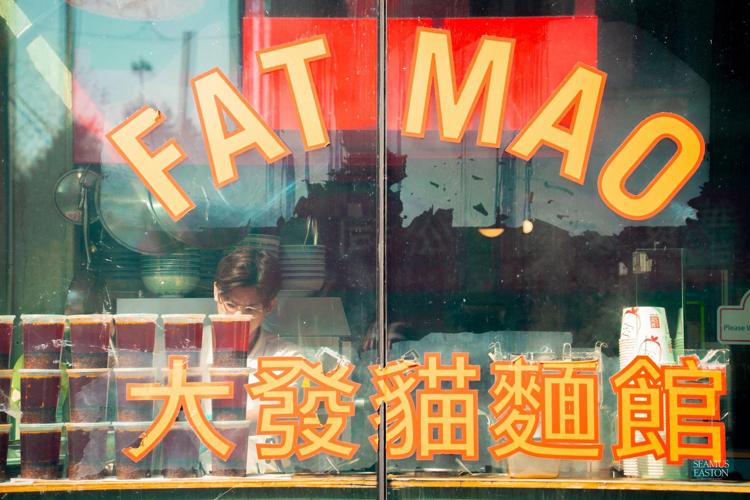 Store window with large orange and yellow letters spelling 'FAT MAO' and Chinese characters underneath, with a person behind the glass preparing drinks and stacked cups inside.