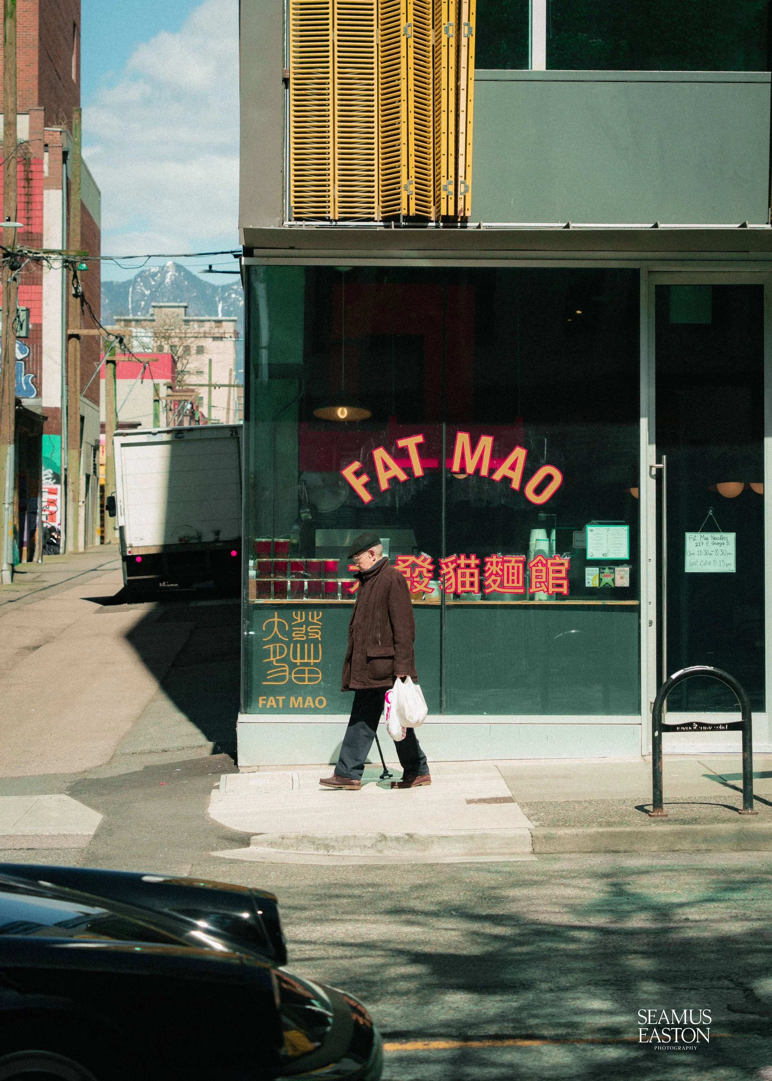 A man walking past a storefront with a sign that reads 'FAT MAO' in large red letters and Chinese characters below it. The person is carrying a bag and wearing a brown coat and black pants, walking on the sidewalk in front of the store with a mountai
