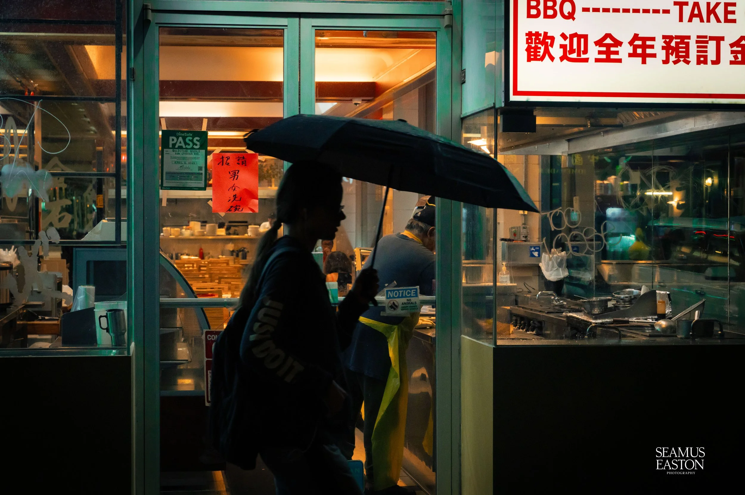 A silhouette of a woman holding an umbrella stands outside a restaurant at night. The restaurant interior is visible through glass, with kitchen staff working. Various signs with Chinese characters and English text are displayed