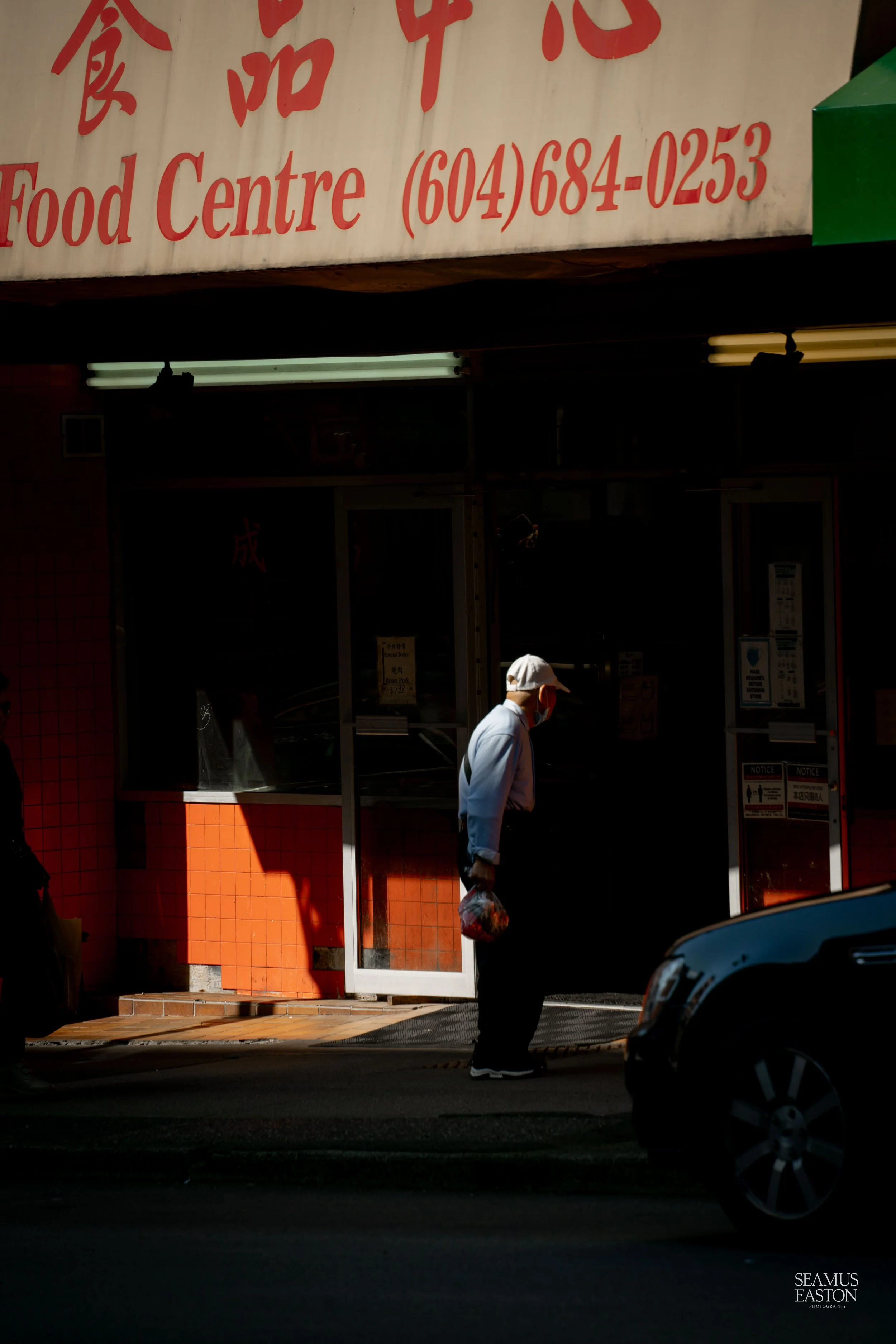 Person wearing a face mask and white cap walking past a Chinese food store with a sign displaying its name, contact number, and some Chinese characters outside.