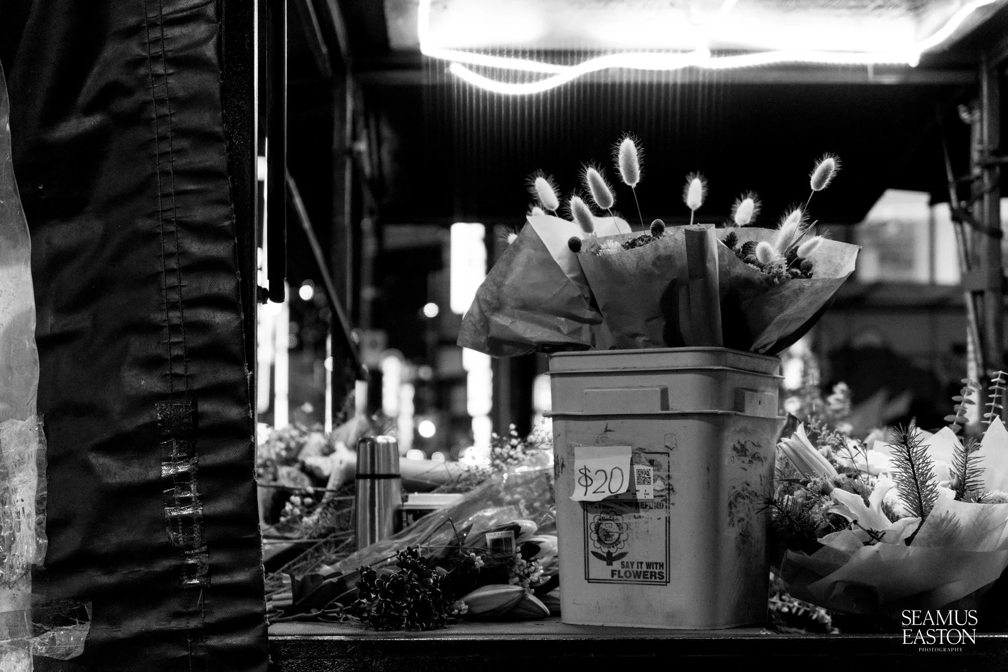 A black and white photo of a flower stall with a box of flowers and various floral arrangements, with a '20' dollar sign price tag attached.