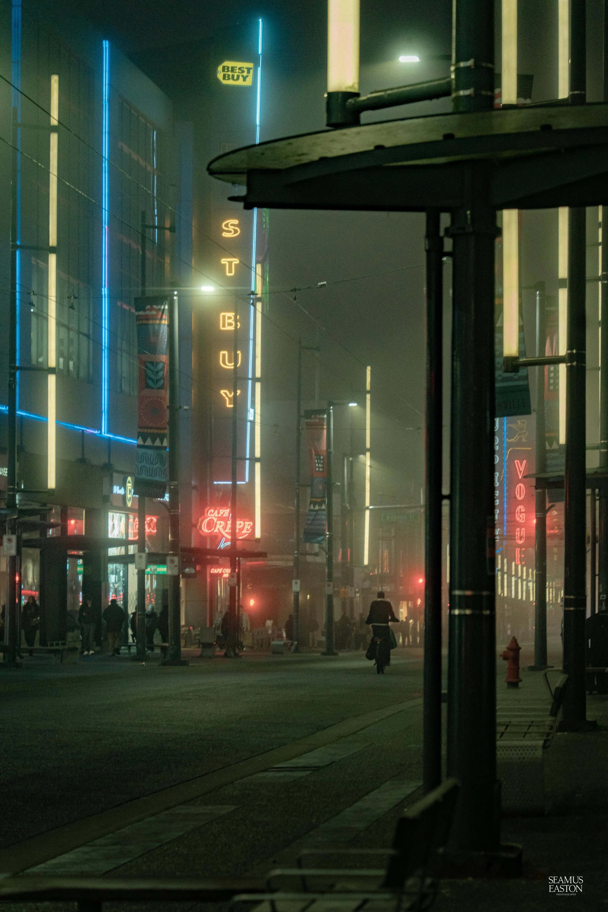 Nighttime city street with neon signs and fog. Signs include 'VOGUE,' 'V,' and 'ST. BUY.' Silhouettes of people walking and a person riding a bicycle are visible. Streetlights and illuminated windows add to the urban atmosphere.