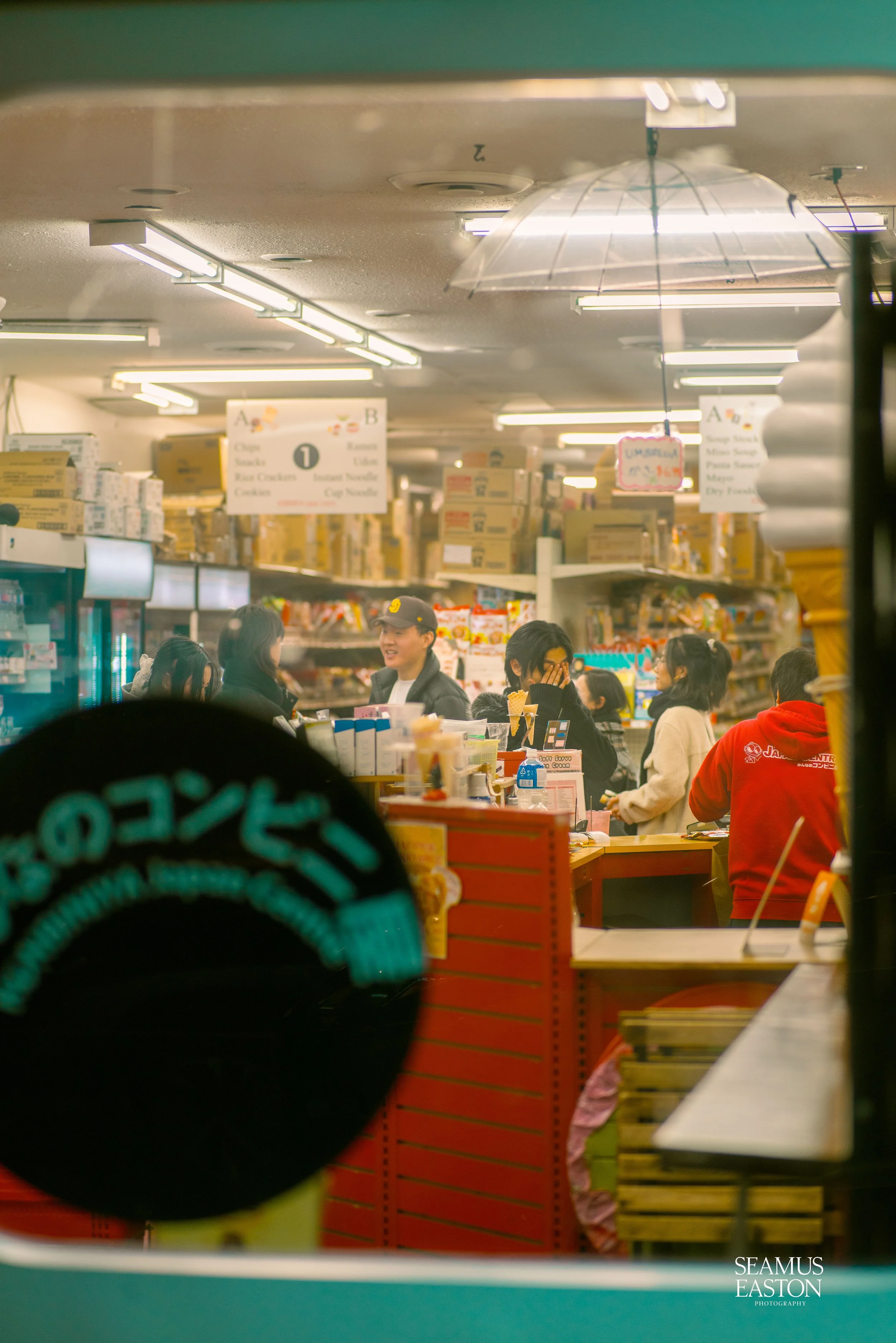 People shopping inside a grocery store viewed through a window with Japanese text and a black circular sign in the foreground.