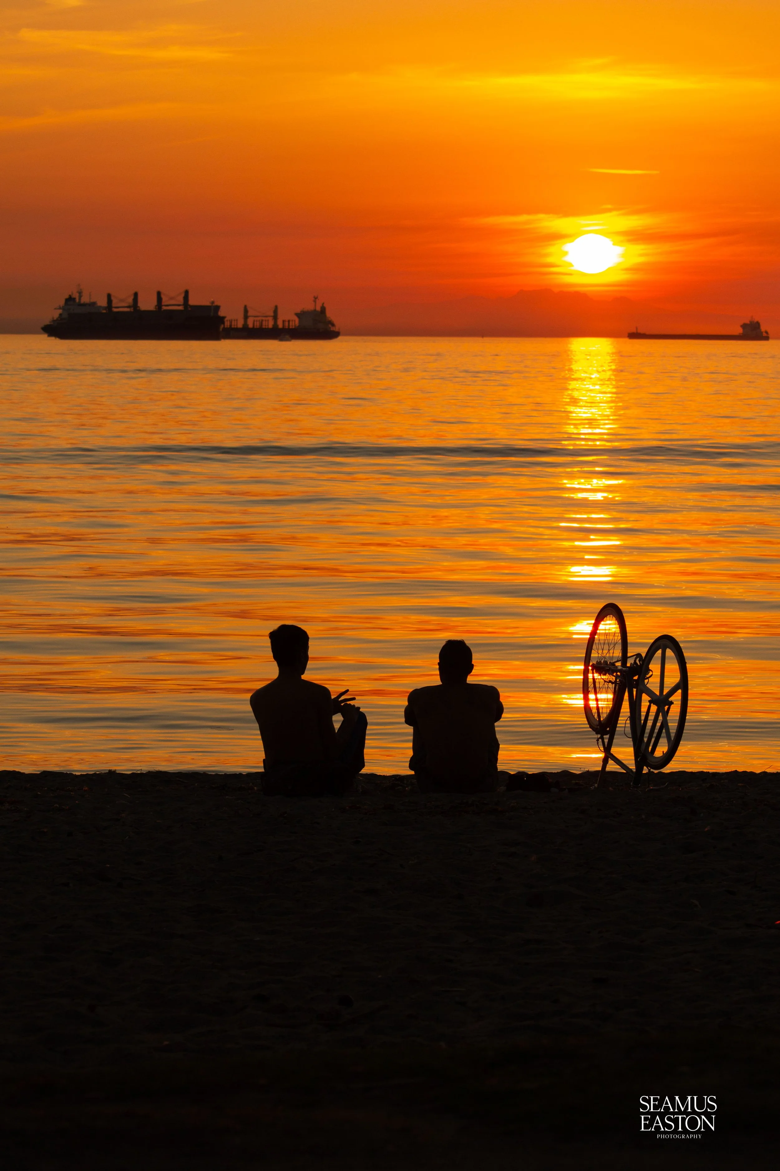 Two people sitting on the beach at sunset, with a bicycle beside them, water with ships, and an orange sky.