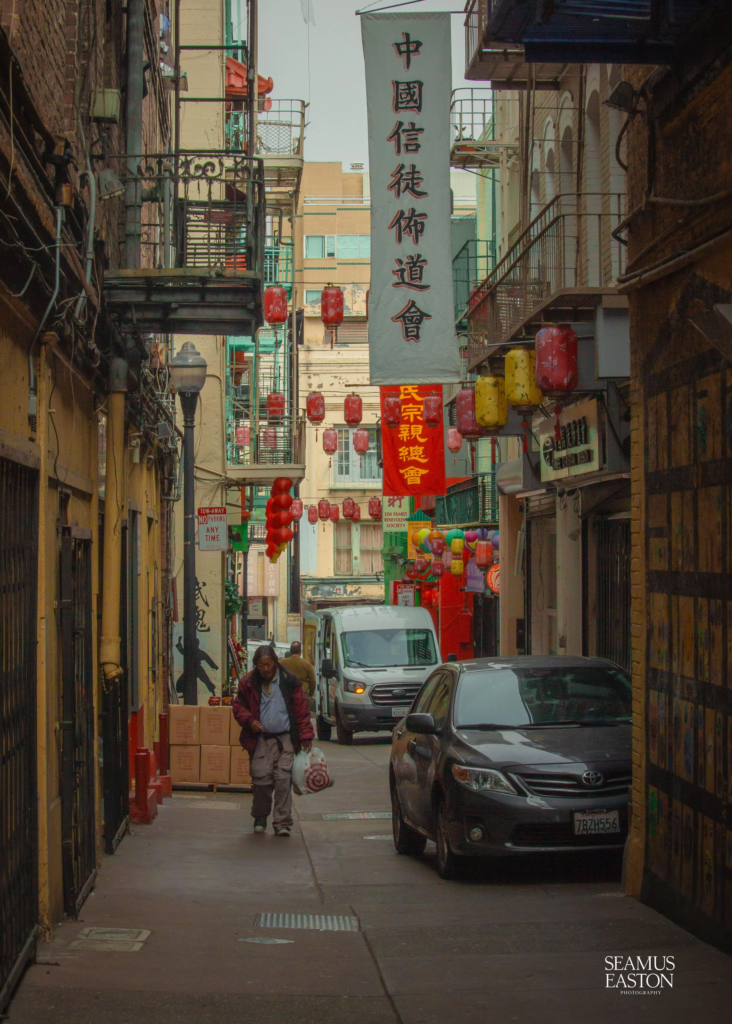 A narrow Chinatown street with hanging red and yellow lanterns, banners with Chinese characters, pedestrians, parked cars, and buildings with fire escapes.