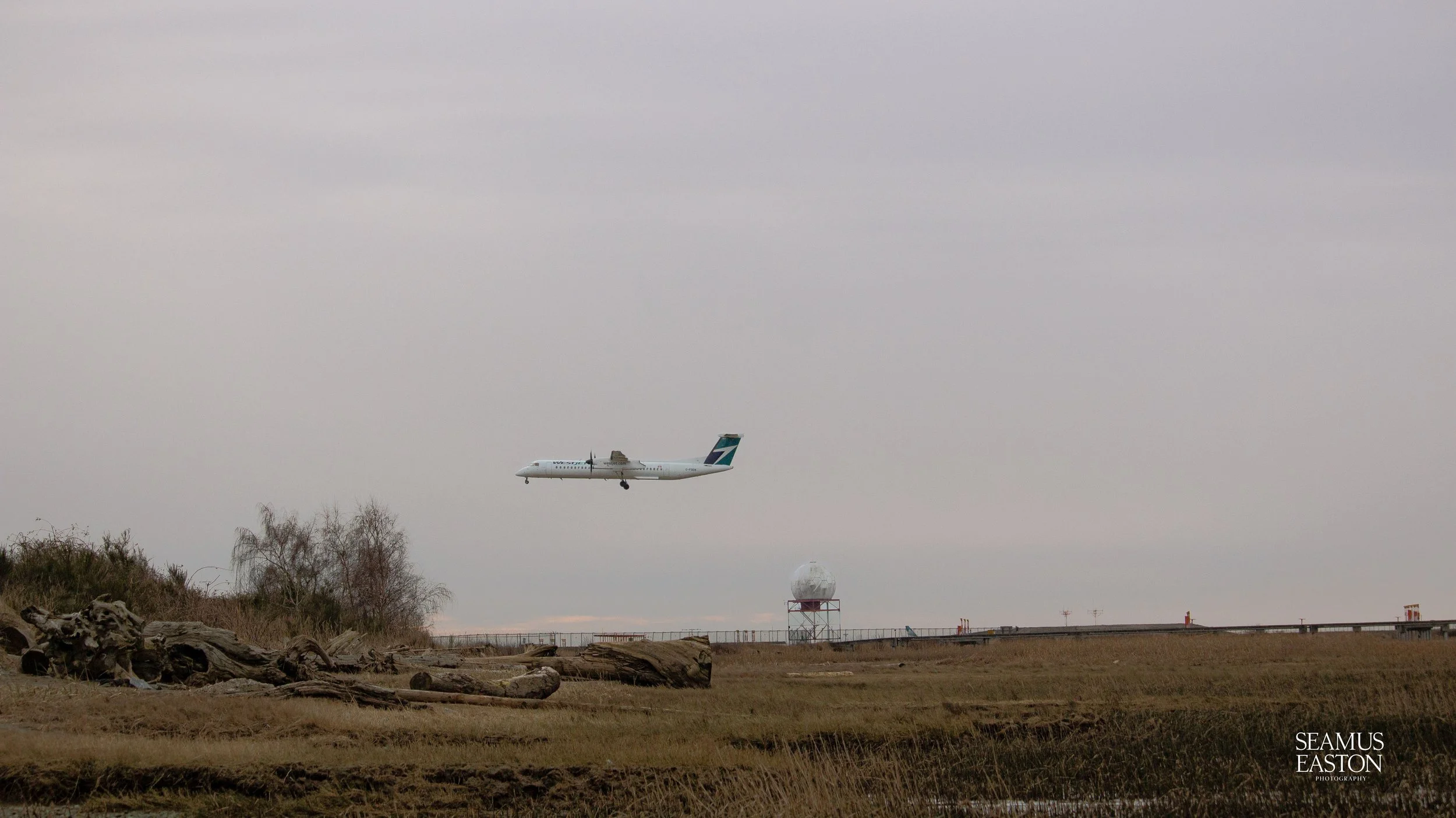 An airplane flying low over a grassy field near a weather radar station on a cloudy day.