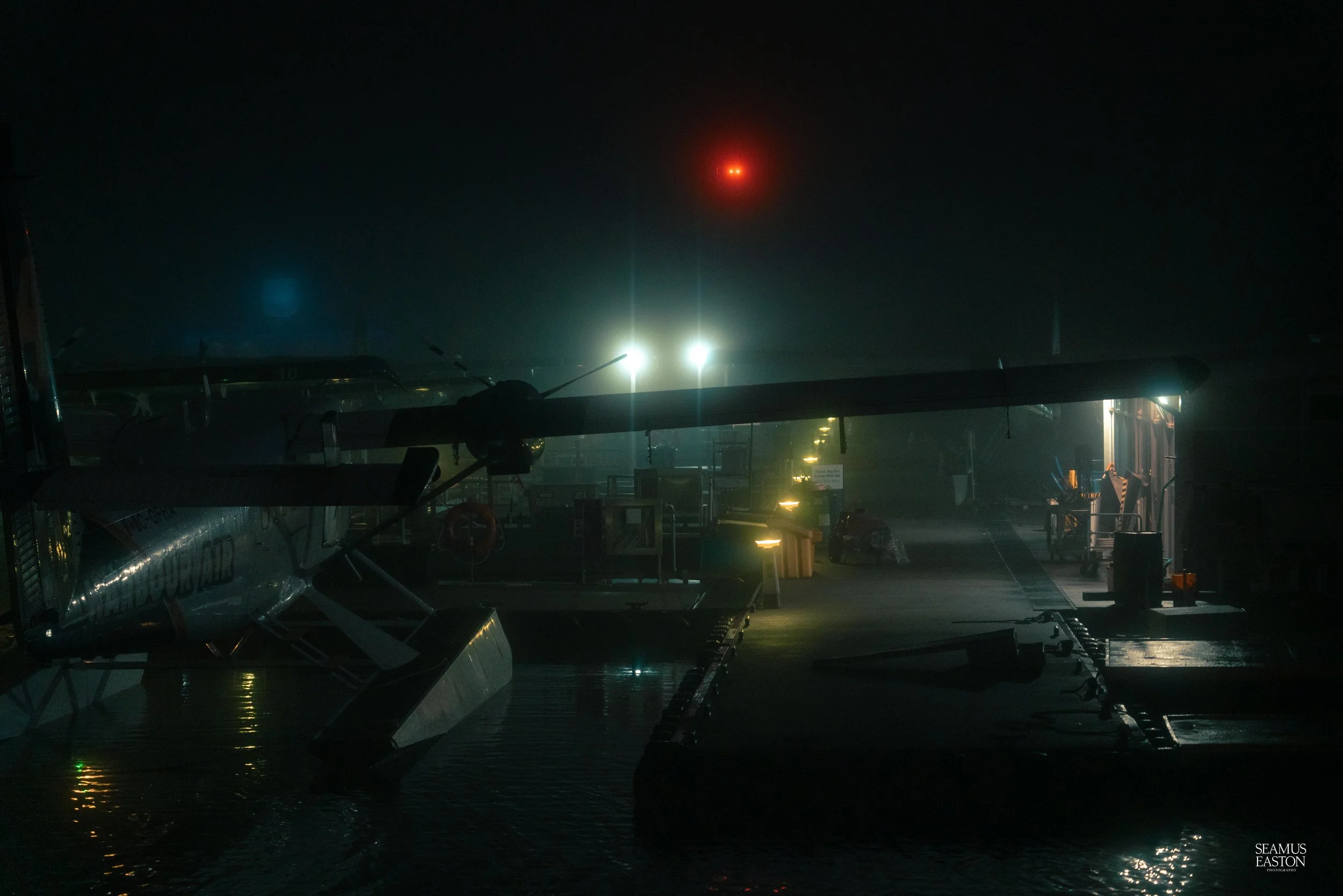 A small seaplane docked at a dark harbor with glowing lights illuminating the scene, night sky in background.
