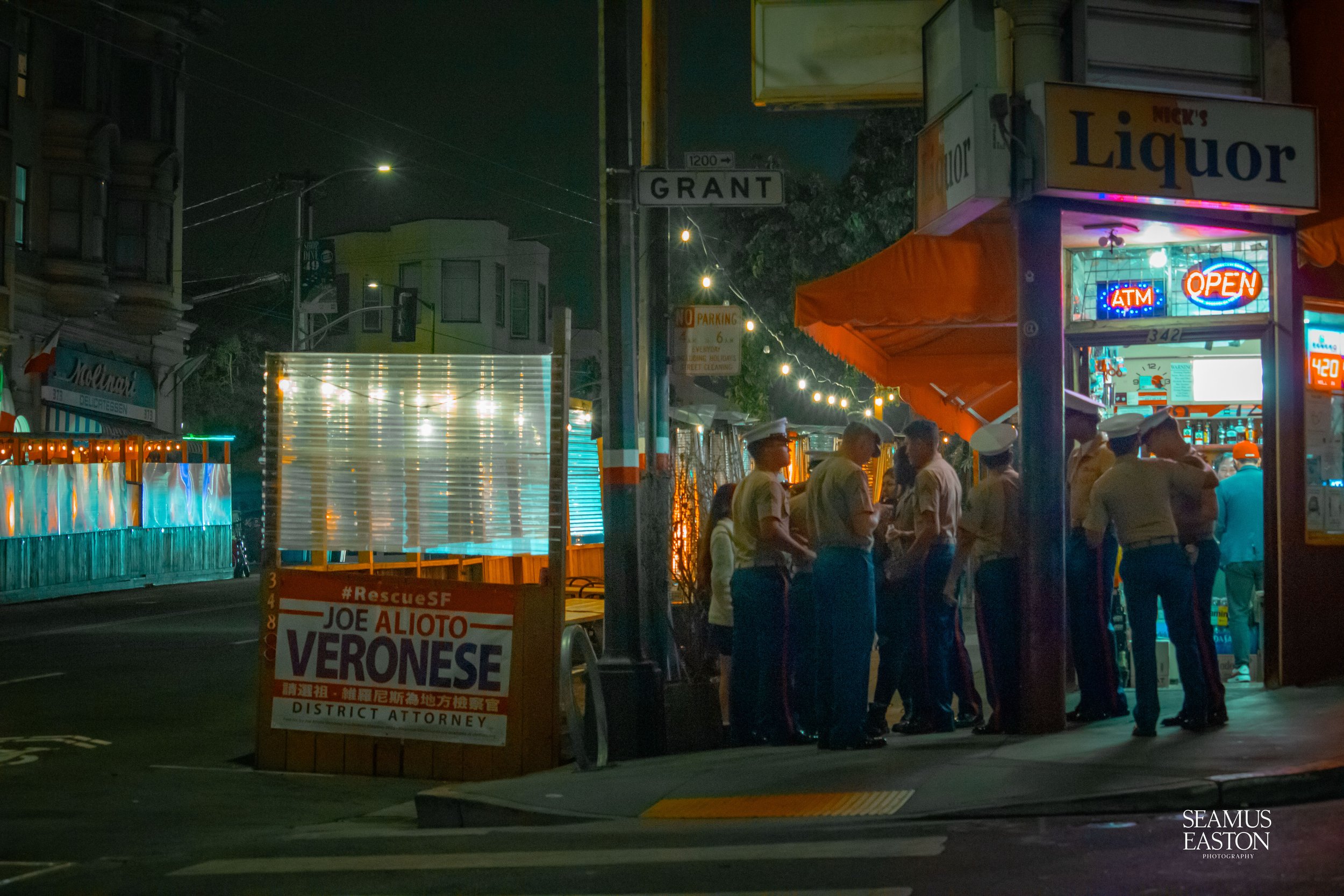 Group of police officers standing outside a liquor store at night, illuminated by neon signs and string lights.