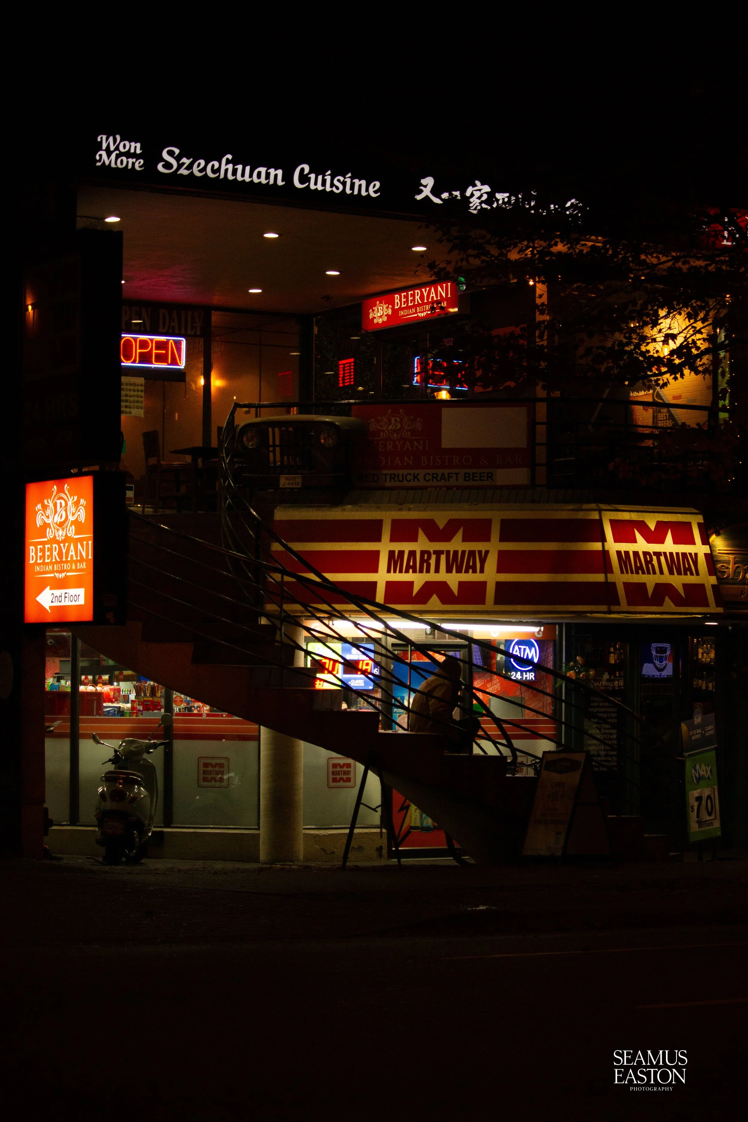 Night view of a restaurant with illuminated signs, including 'Szechuan Cuisine' and 'Beeryani Indian Bistro & Bar', and a person sitting on the stairs outside.
