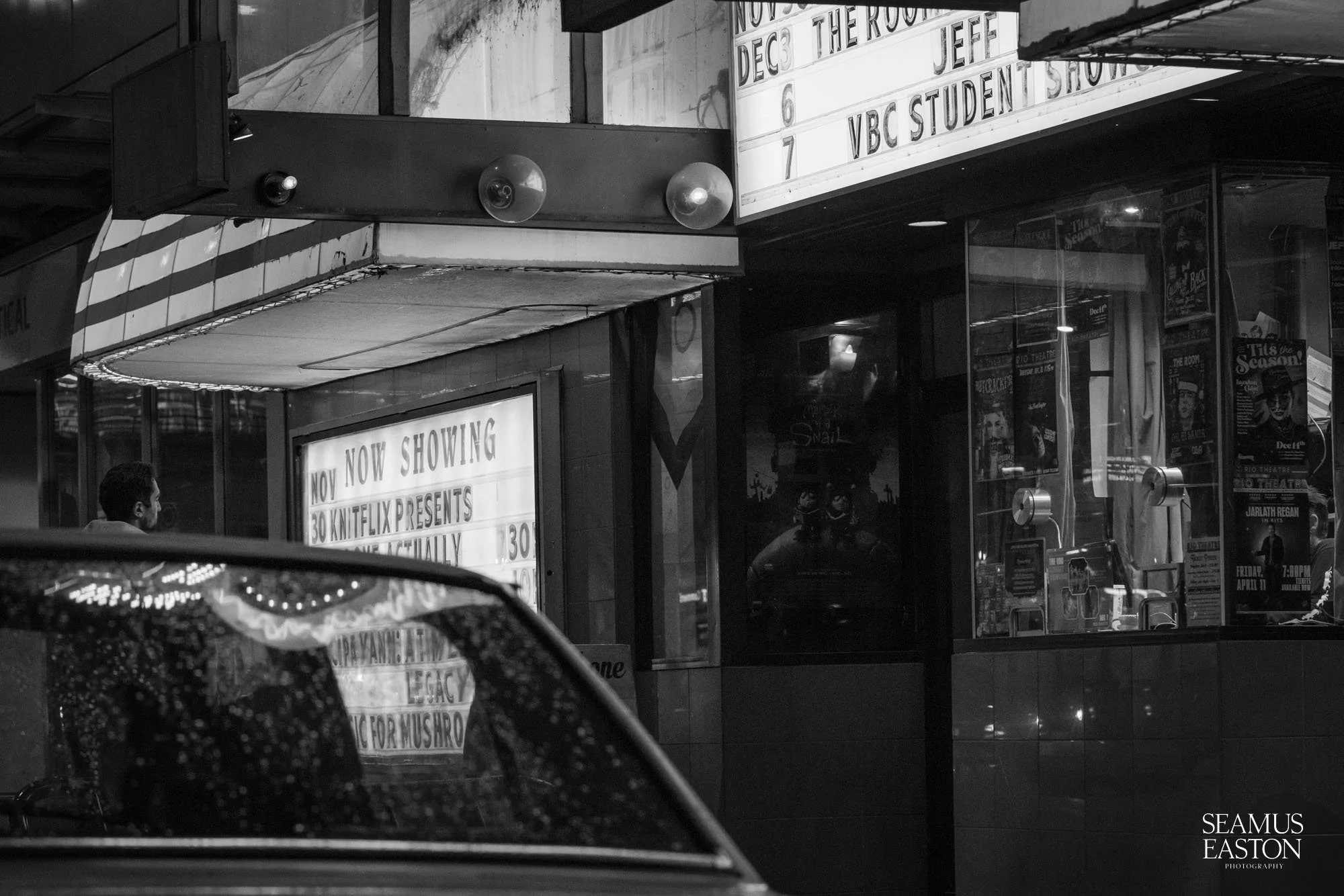 Night view of a city street corner showing a theater marquee with movie listings, a person standing nearby, and a car with rain droplets on its windshield in the foreground.