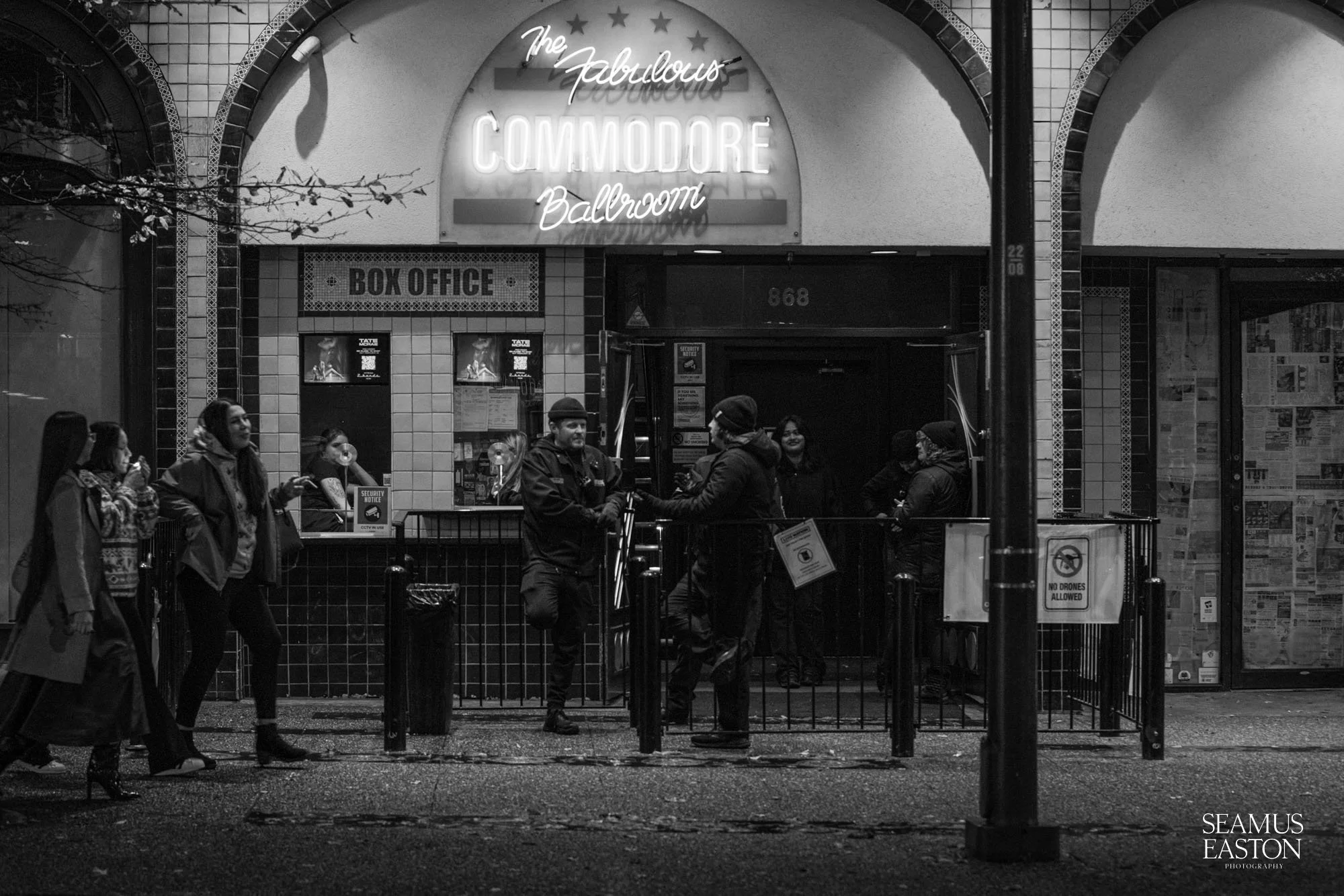 People standing outside the entrance of The Fabulous Commodore Ballroom, with some in line and others talking, during evening or night.