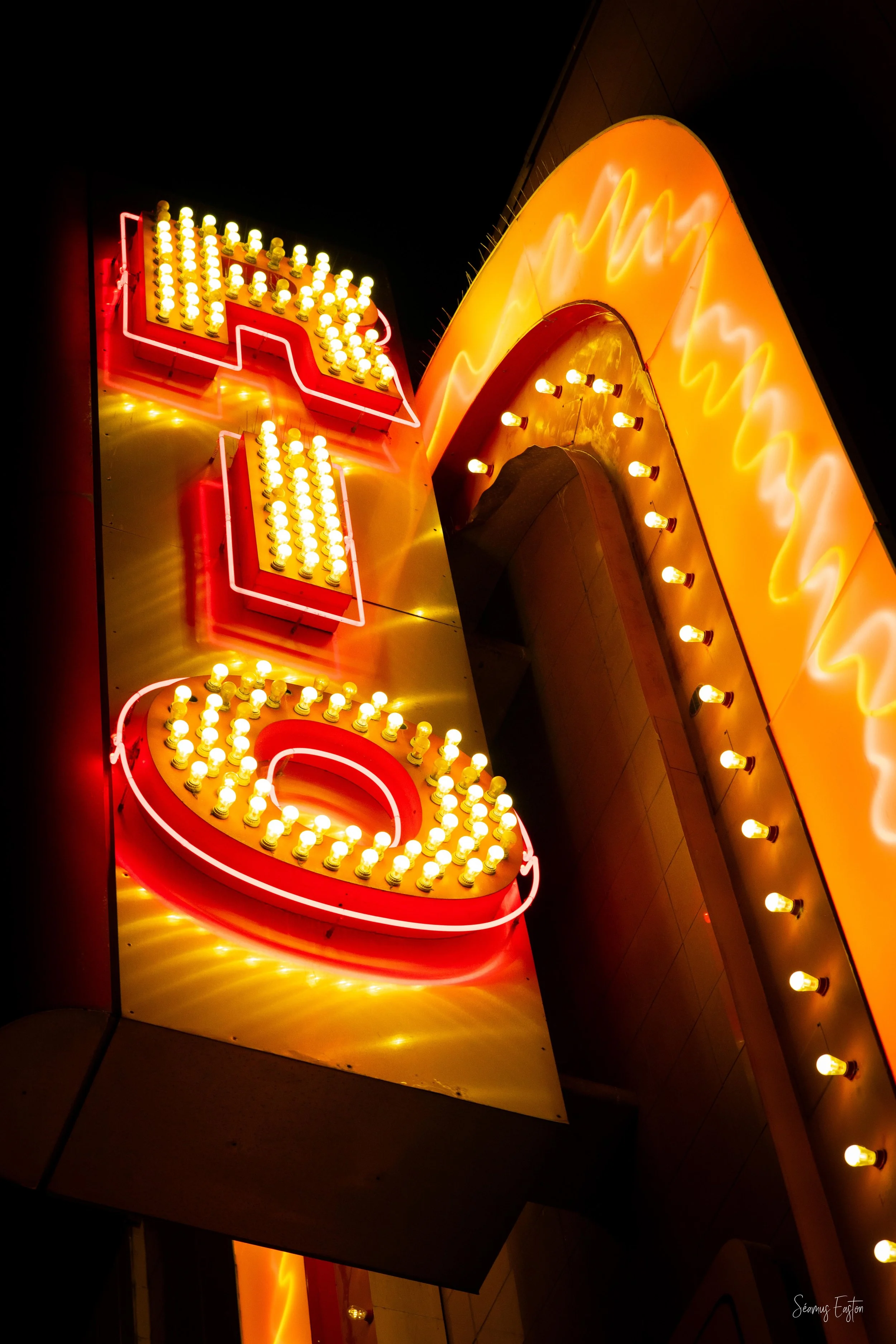 Close-up of a bright vintage neon sign with the word "HOTEL," featuring yellow and red light bulbs against a dark night background.