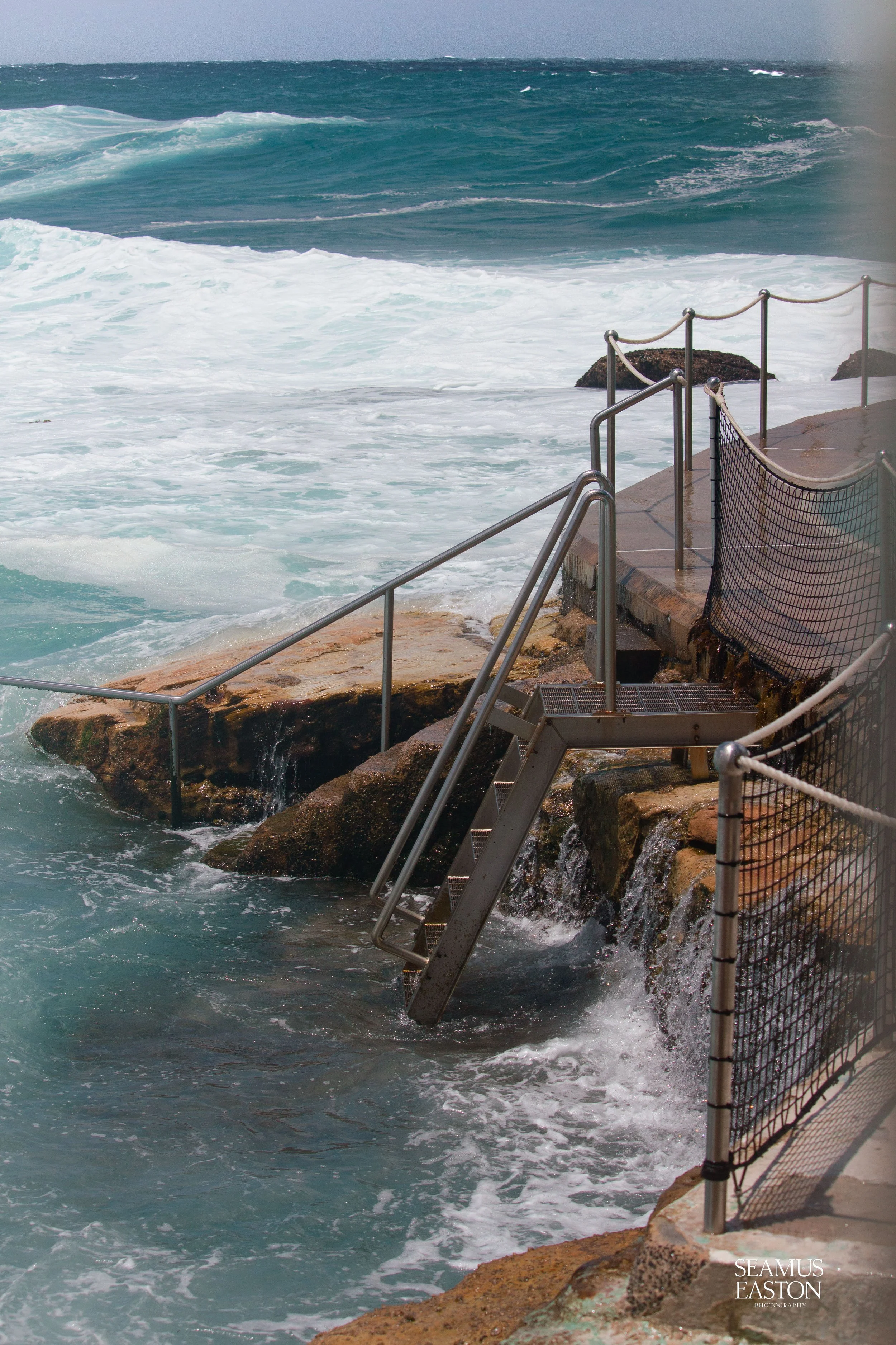Ocean waves crash against a rocky shoreline with metal stairs and safety railing leading into the water at a beach.