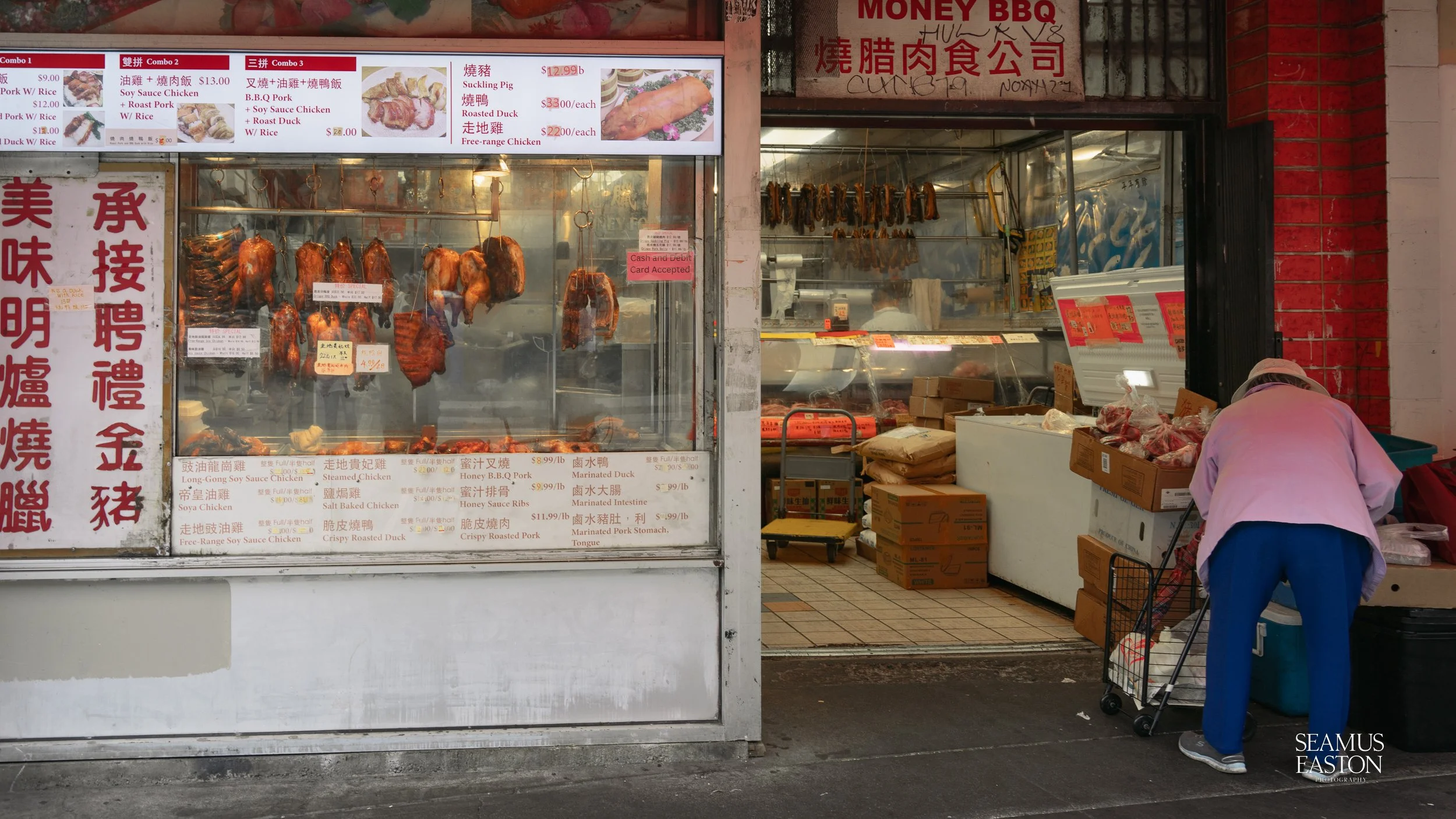 A street food stall with hanging roasted ducks and pigs, with menus in English and Chinese, and a person shopping for meat.