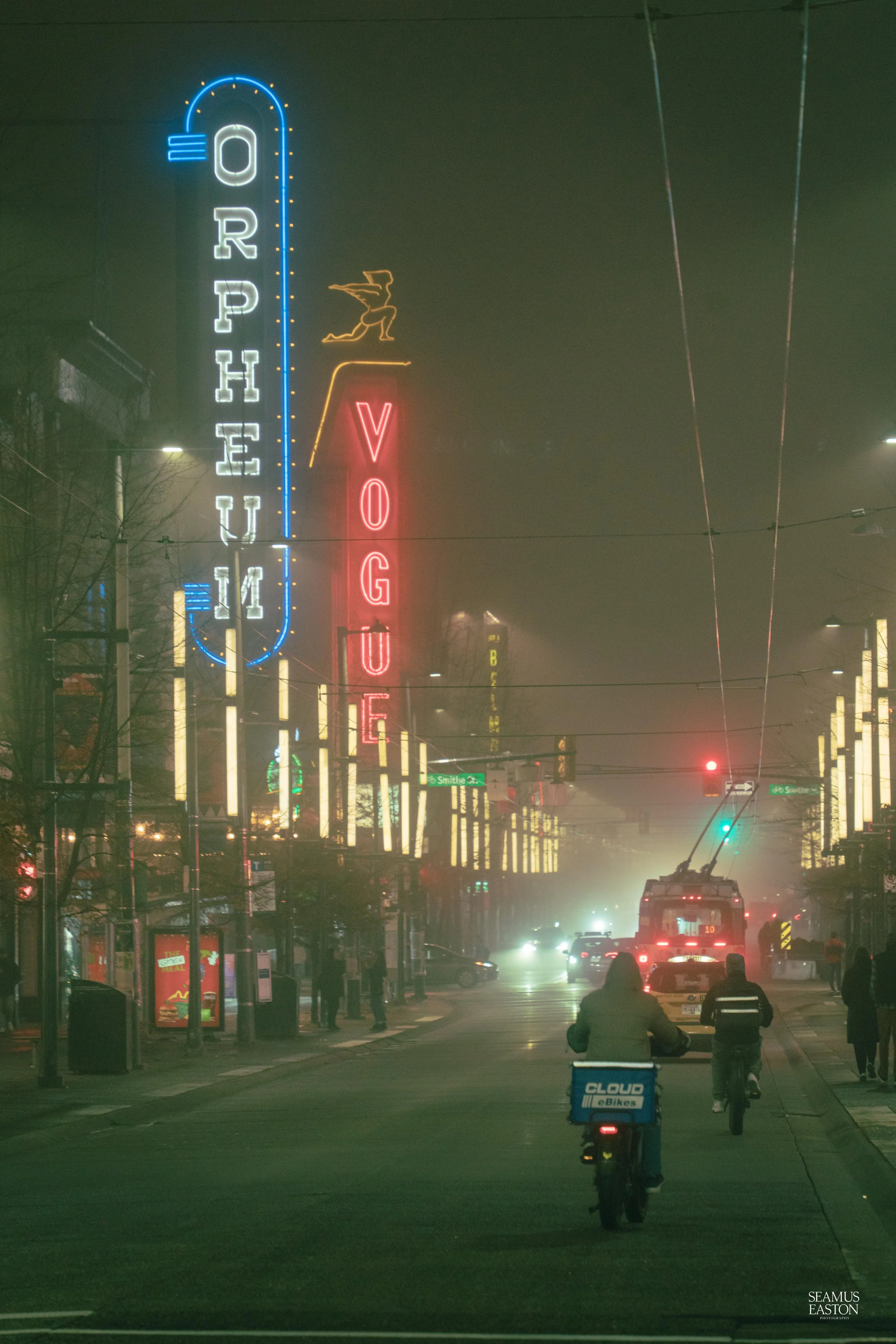 Night street scene with neon signs, including a large blue and white "OPHEU 14" sign, a red "VOGUE" sign, and a smaller illuminated witch figure on top. Pedestrians and cyclists on the street, with cars and an streetlight in the background. Foggy atm