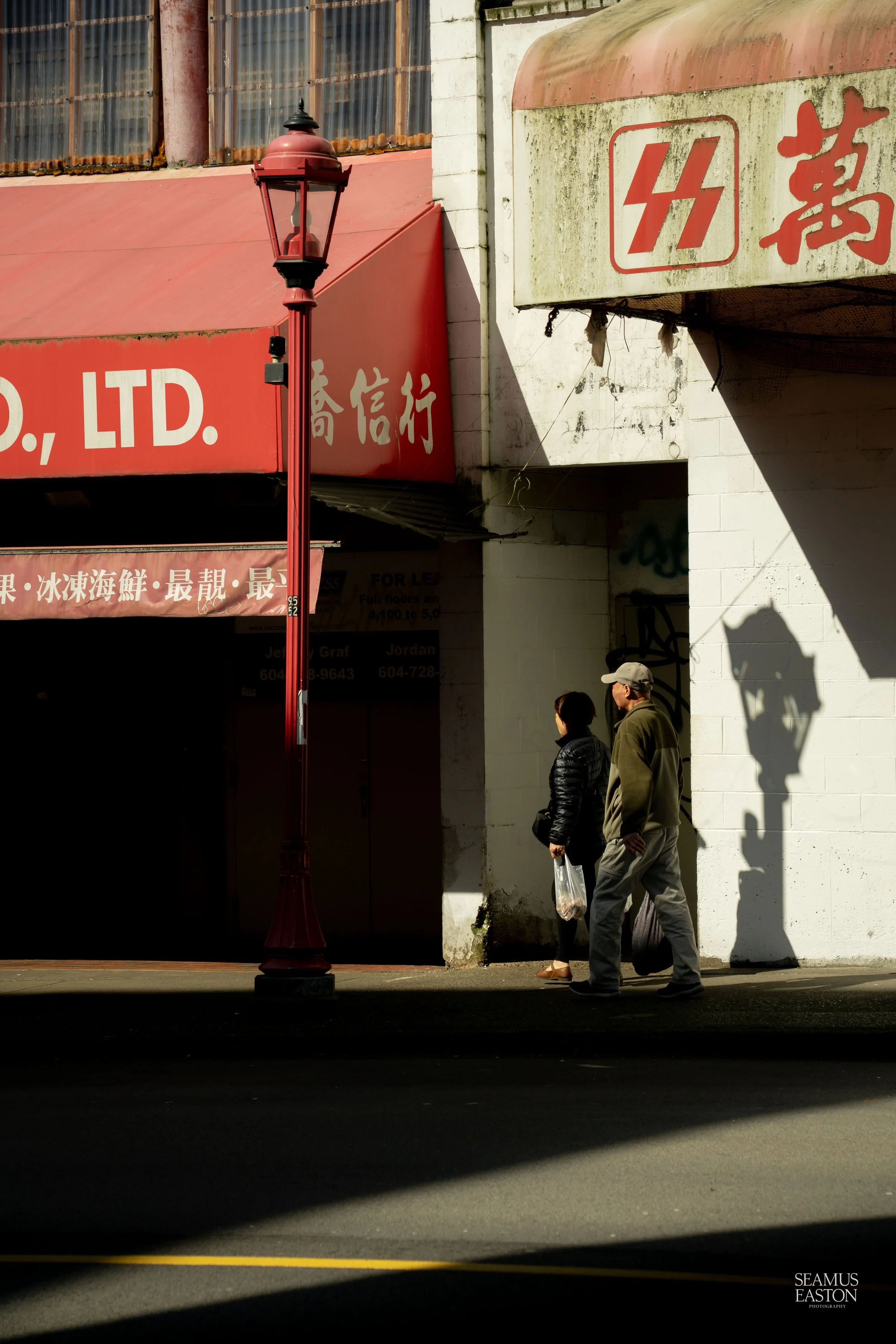 A man and woman walking on a city sidewalk past a red street lamp, with Asian signs and storefronts, and shadows cast on white building walls.