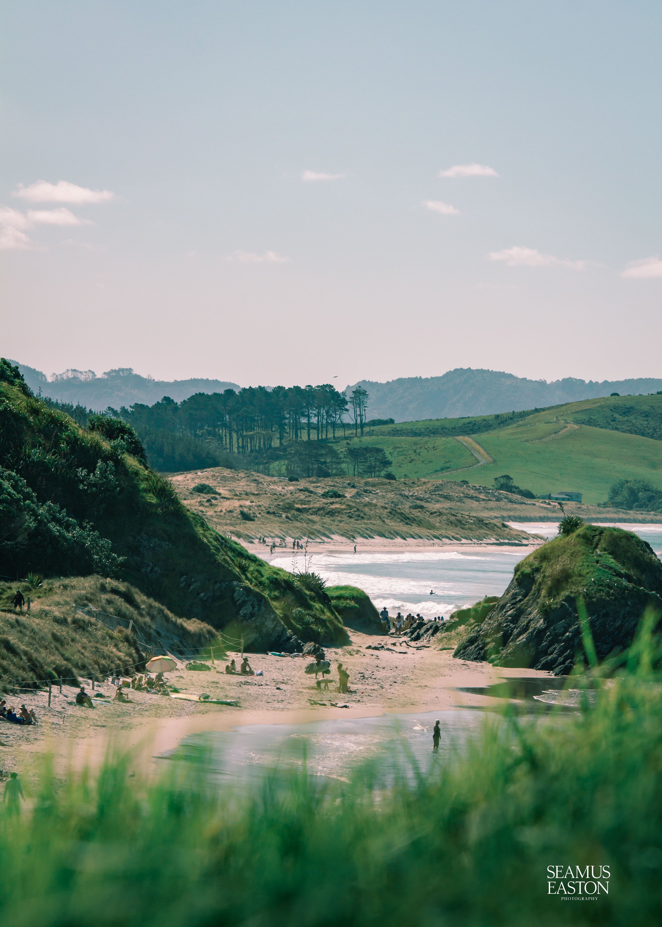 A beach with green hills on either side, a sandy shoreline with people, and distant green mountains under a partly cloudy sky.