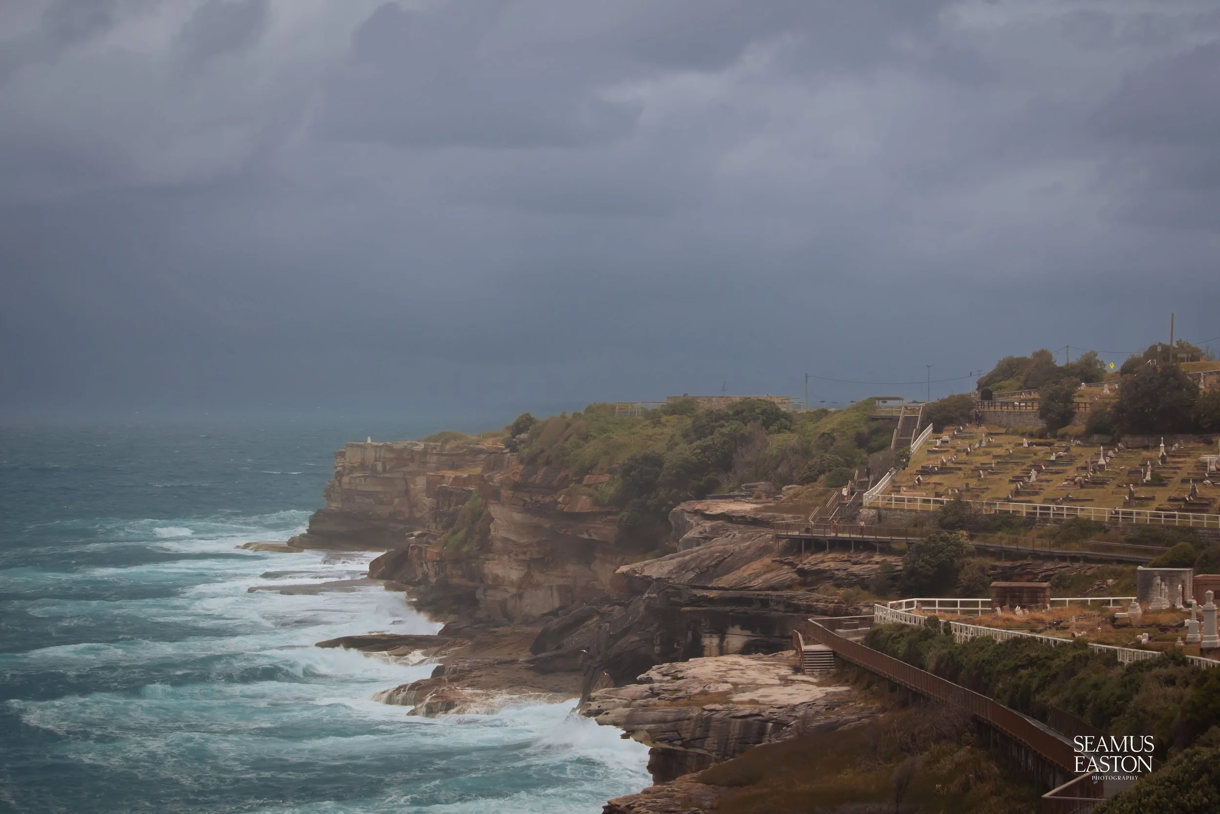 Cliffs and rocky shoreline with ocean waves under cloudy sky, beside a cemetery with many gravestones and shrubs, near the coast.