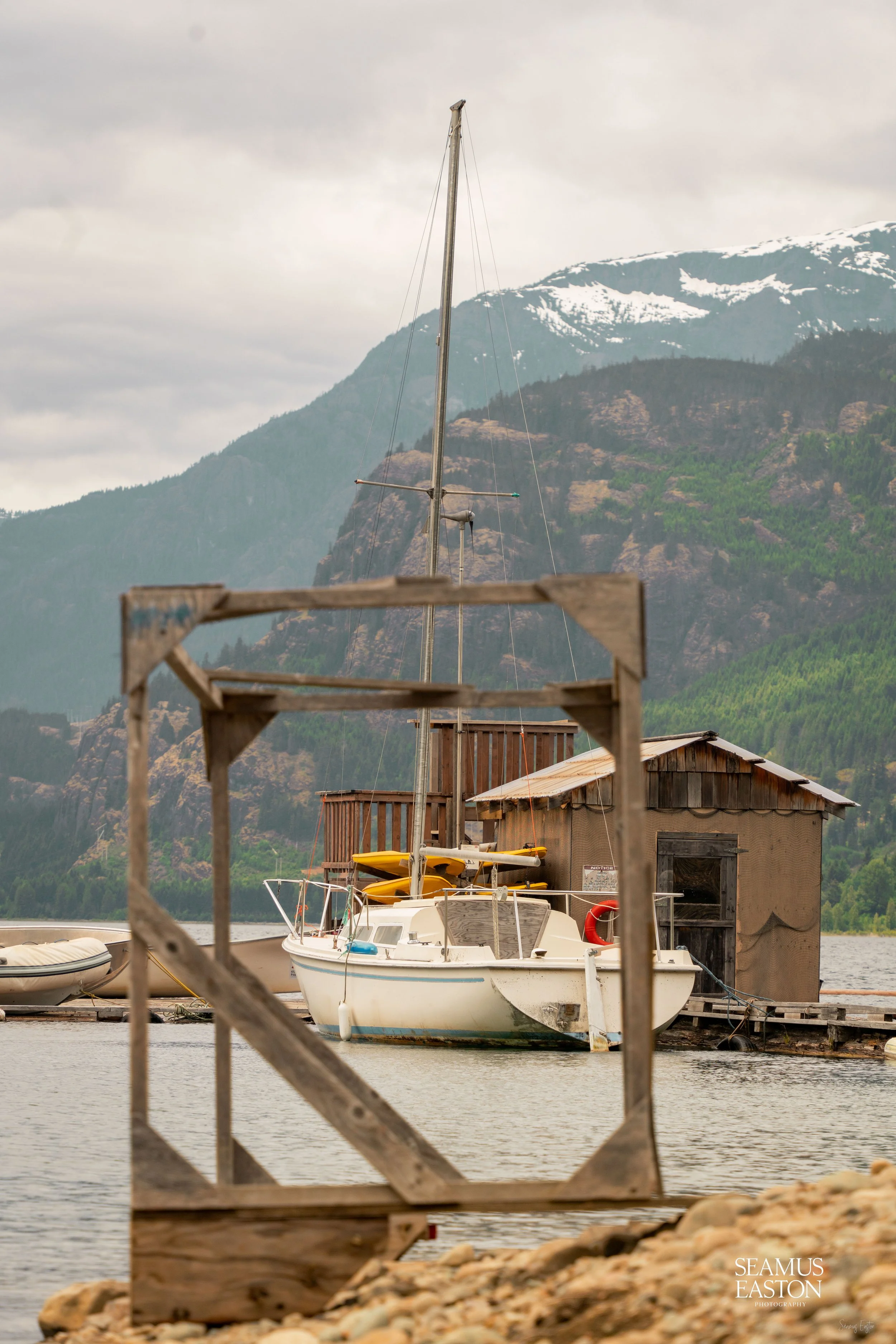 A sailboat docked by a wooden boathouse on a lake, with a mountainous landscape in the background.