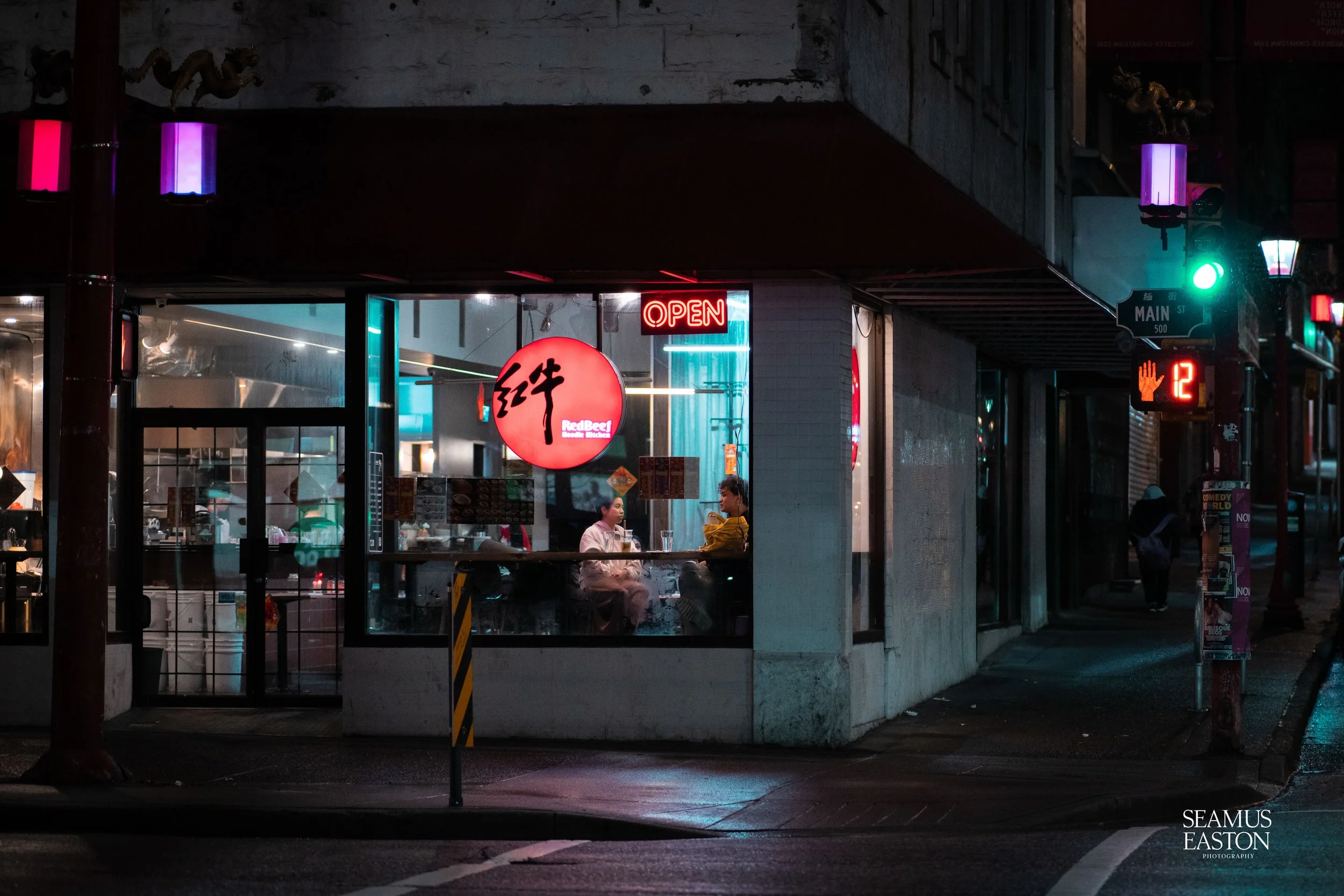 Nighttime street scene with a restaurant window illuminated by neon signs, including an 'OPEN' sign and a red circular sign with Asian characters and 'Red Beef' text, showing two people sitting inside at a table engaged in conversation.