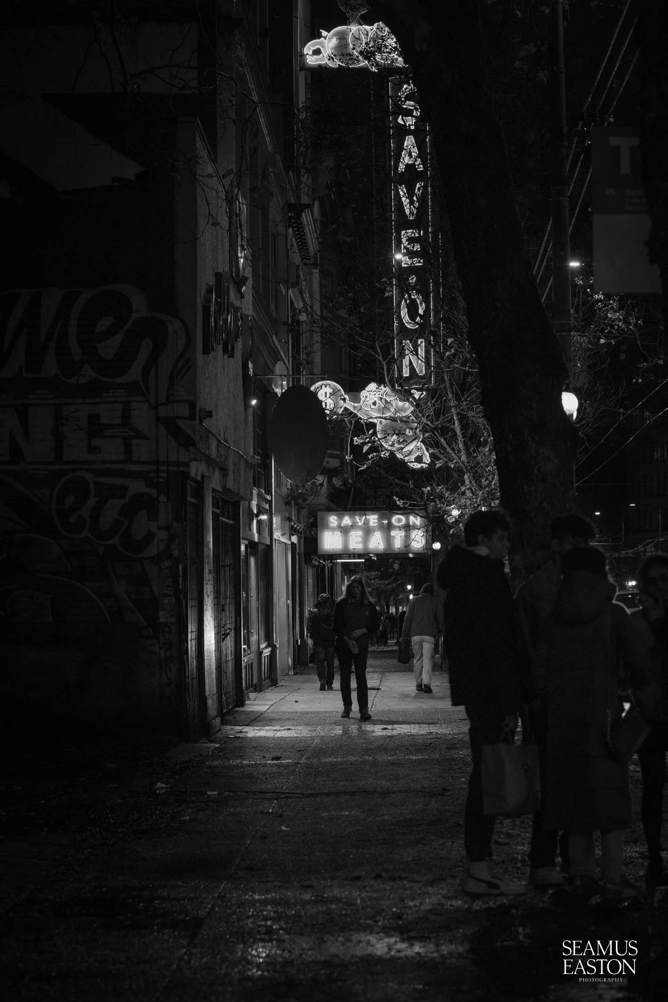 A black and white photo of a city sidewalk at night with illuminated signs, including a vertical neon sign reading 'TAVERN' and a horizontal sign saying 'SAVE ON MEATS.' Several people are walking and standing along the sidewalk, with some visible in