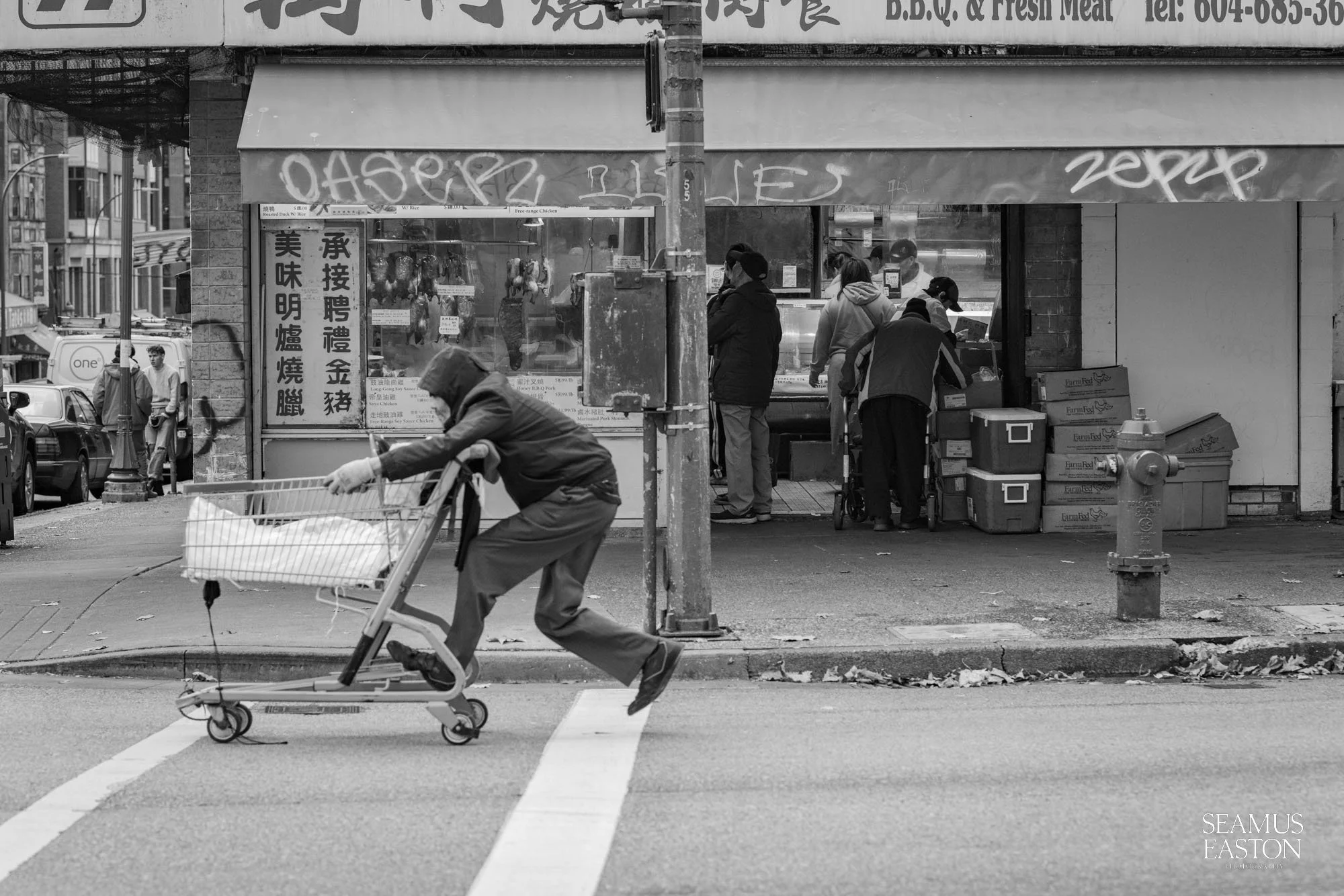 A person crossing the street with a shopping cart in front of a storefront with people inside and graffiti on the awning in an urban setting