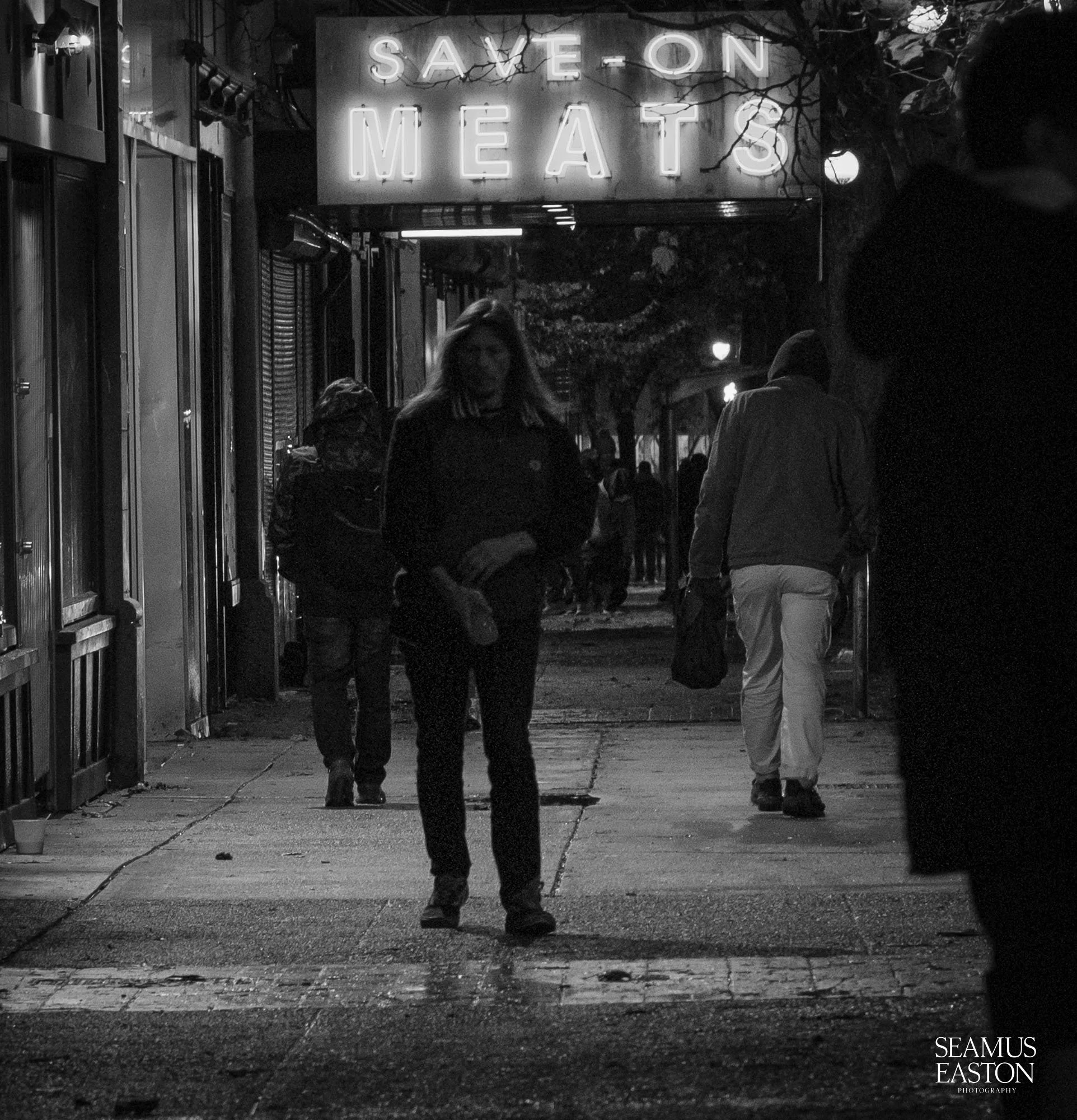 Night scene of a city sidewalk with several people walking; illuminated sign overhead reads 'Save on Meats'.