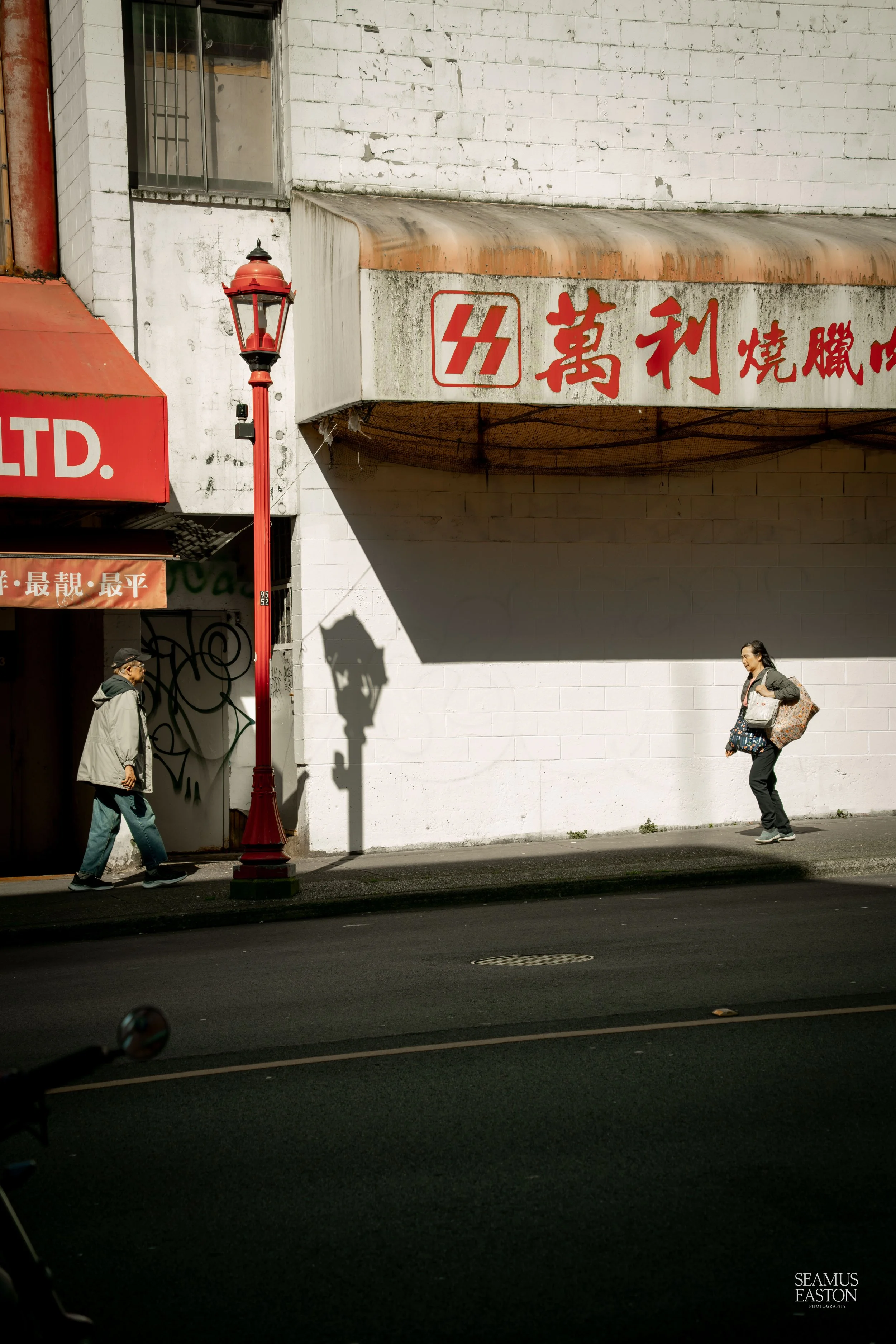 Two people walking on a city sidewalk next to a white wall with red Chinese characters and a sign with a red logo. The scene includes a red street lamp casting a shadow on the wall, with a building featuring graffiti and a small window in the backgro