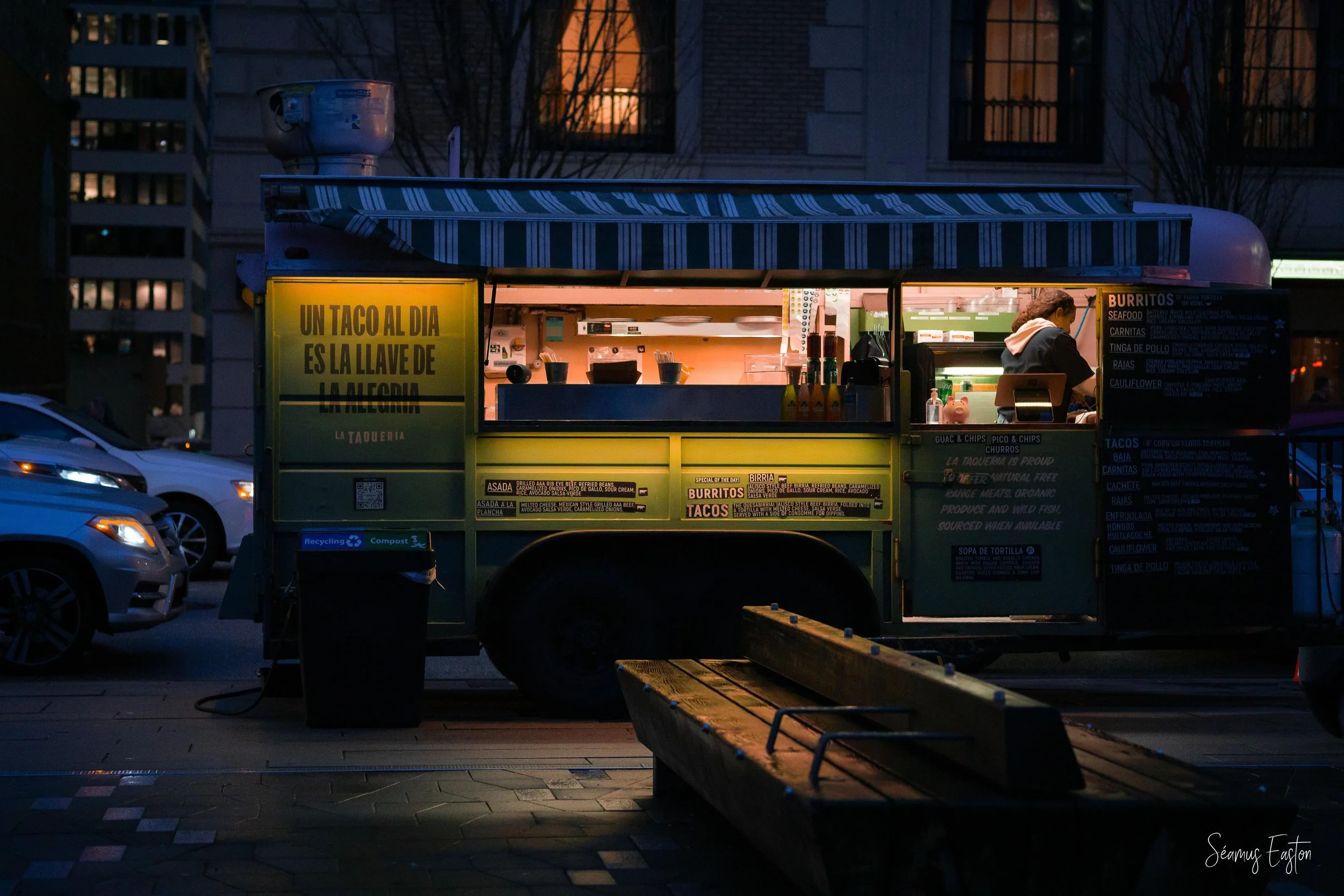 A street food truck illuminated at dusk, selling tacos and burritos in an urban area with parked cars nearby.