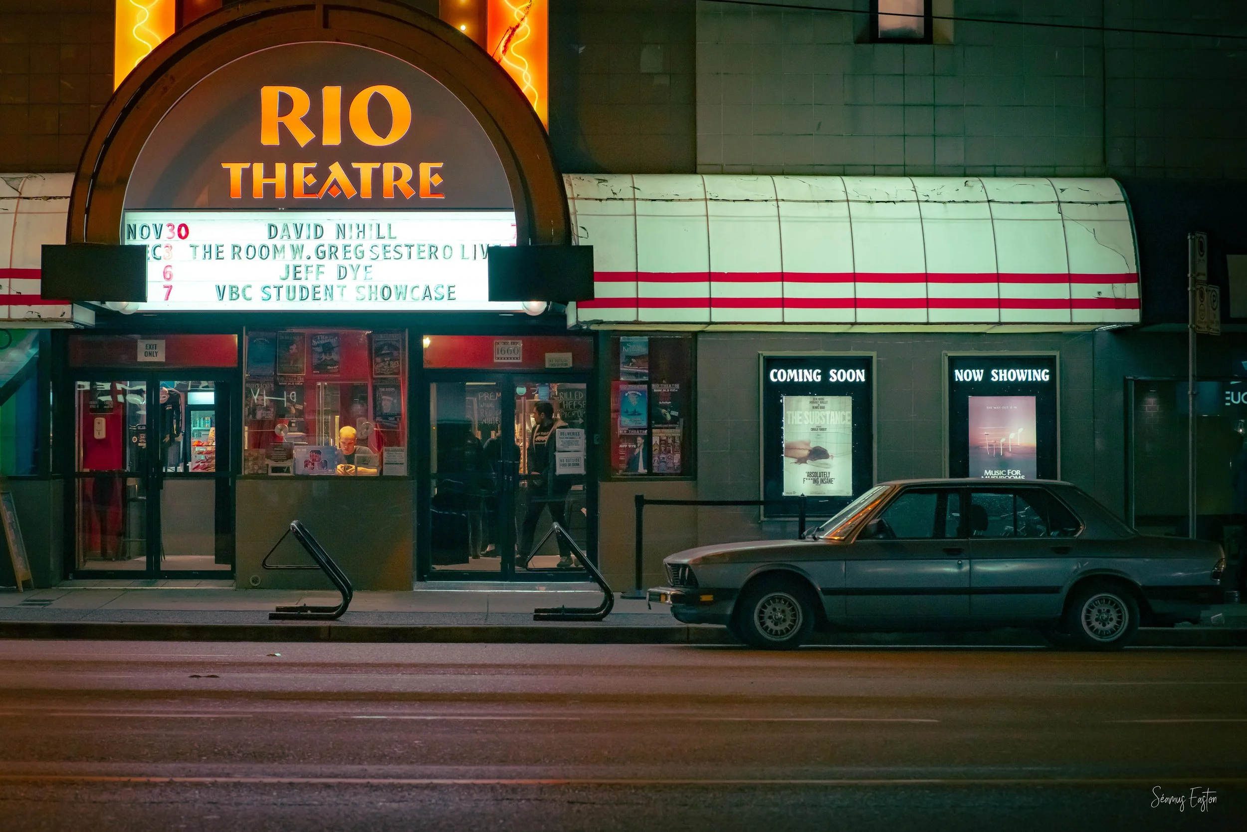 Exterior view of Rio Theatre at night with illuminated marquee sign, showing upcoming shows, a vintage car parked in front, and posters advertising movies 'Coming Soon' and 'Now Showing'.