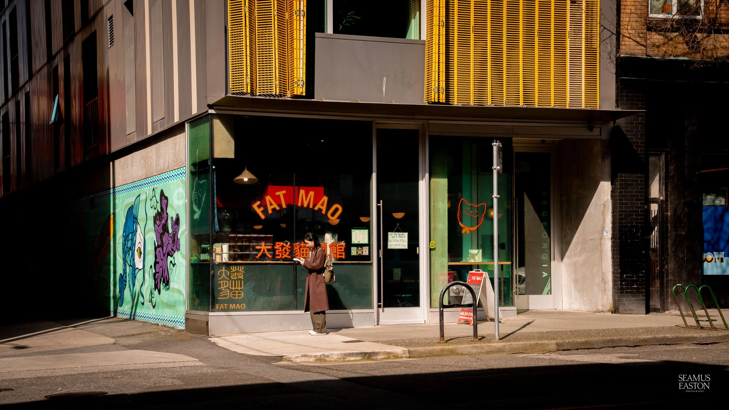 A woman in a brown coat and face mask reads a menu outside a restaurant called FAT MAO, with graffiti art of a fish and purple ink on the building's wall. Sunlight creates shadows on the sidewalk.