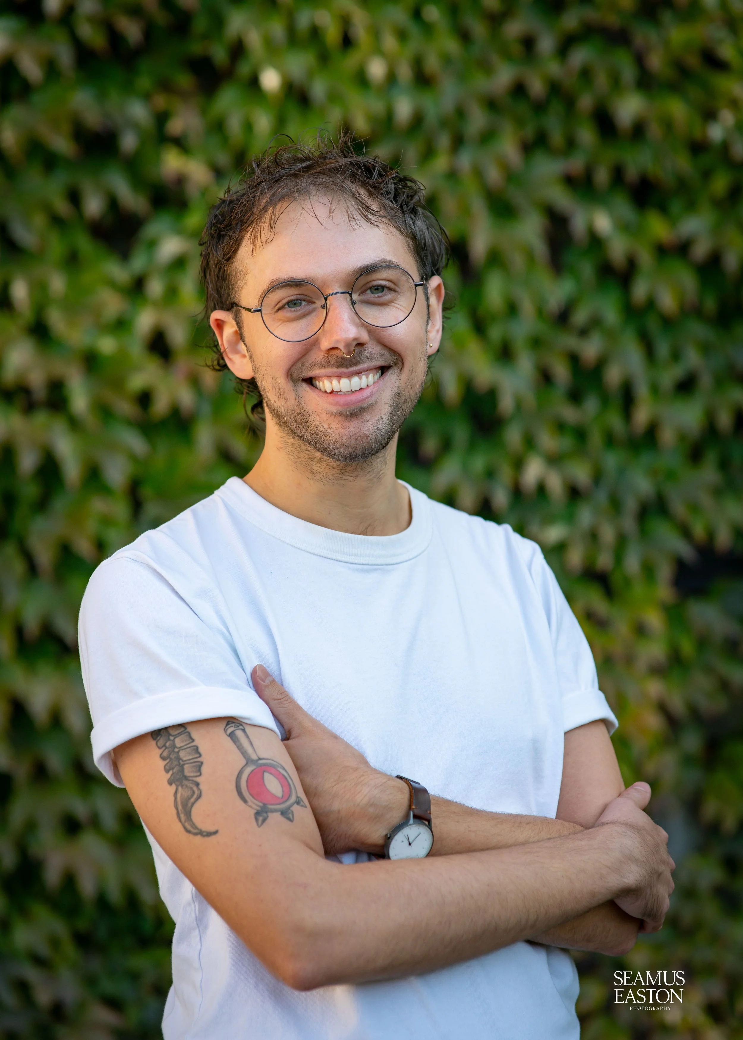 Young man with glasses and tattoos on his arm smiling with arms crossed outdoors in front of a green leafy background.