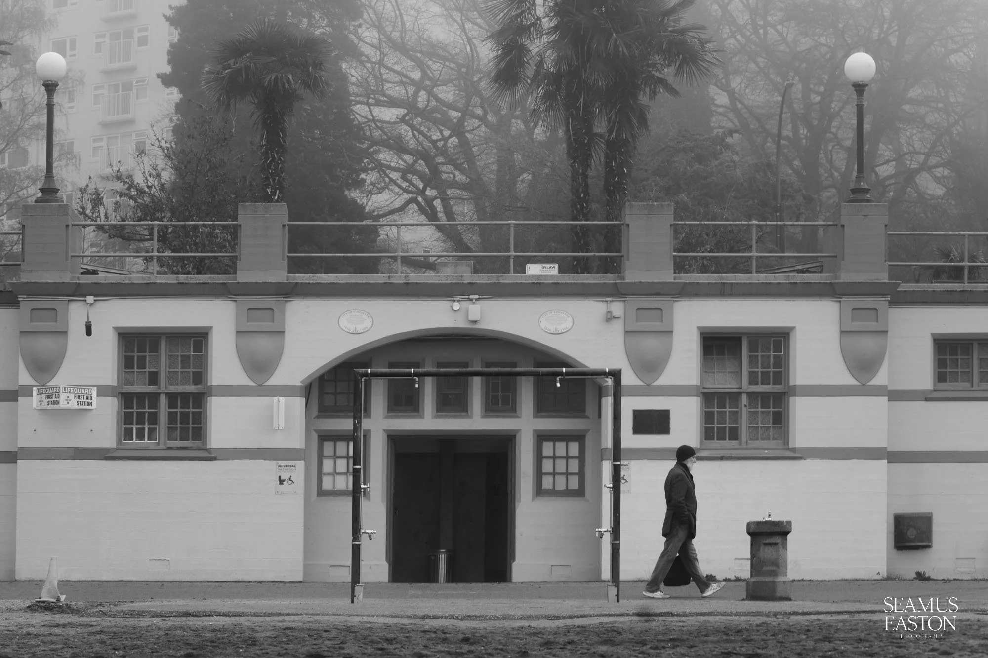 A person walking past an old building with arched entrance and decorative details on the facade, surrounded by trees in foggy weather.