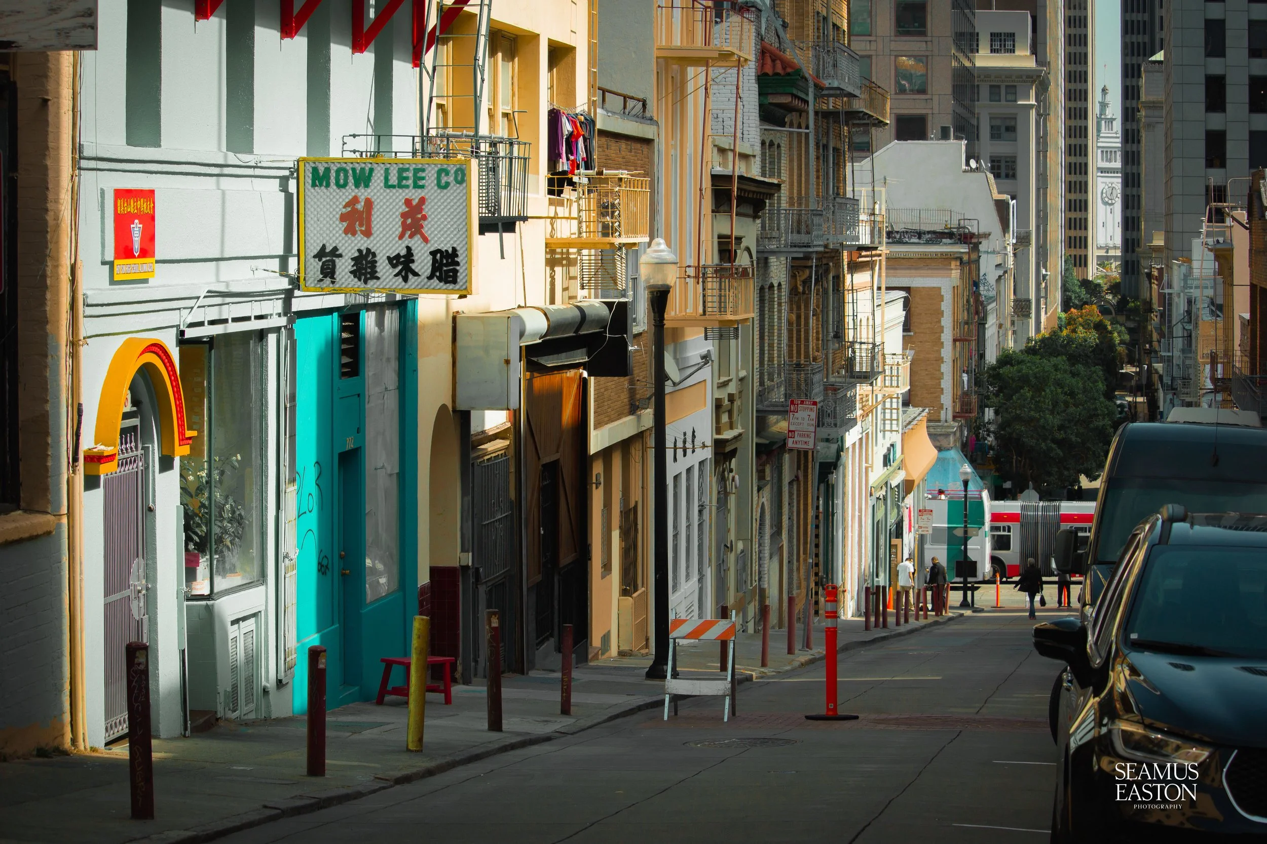 A city street scene with colorful storefronts, security posts, street poles, a lamppost, pedestrians, and parked cars. High-rise buildings are in the background.