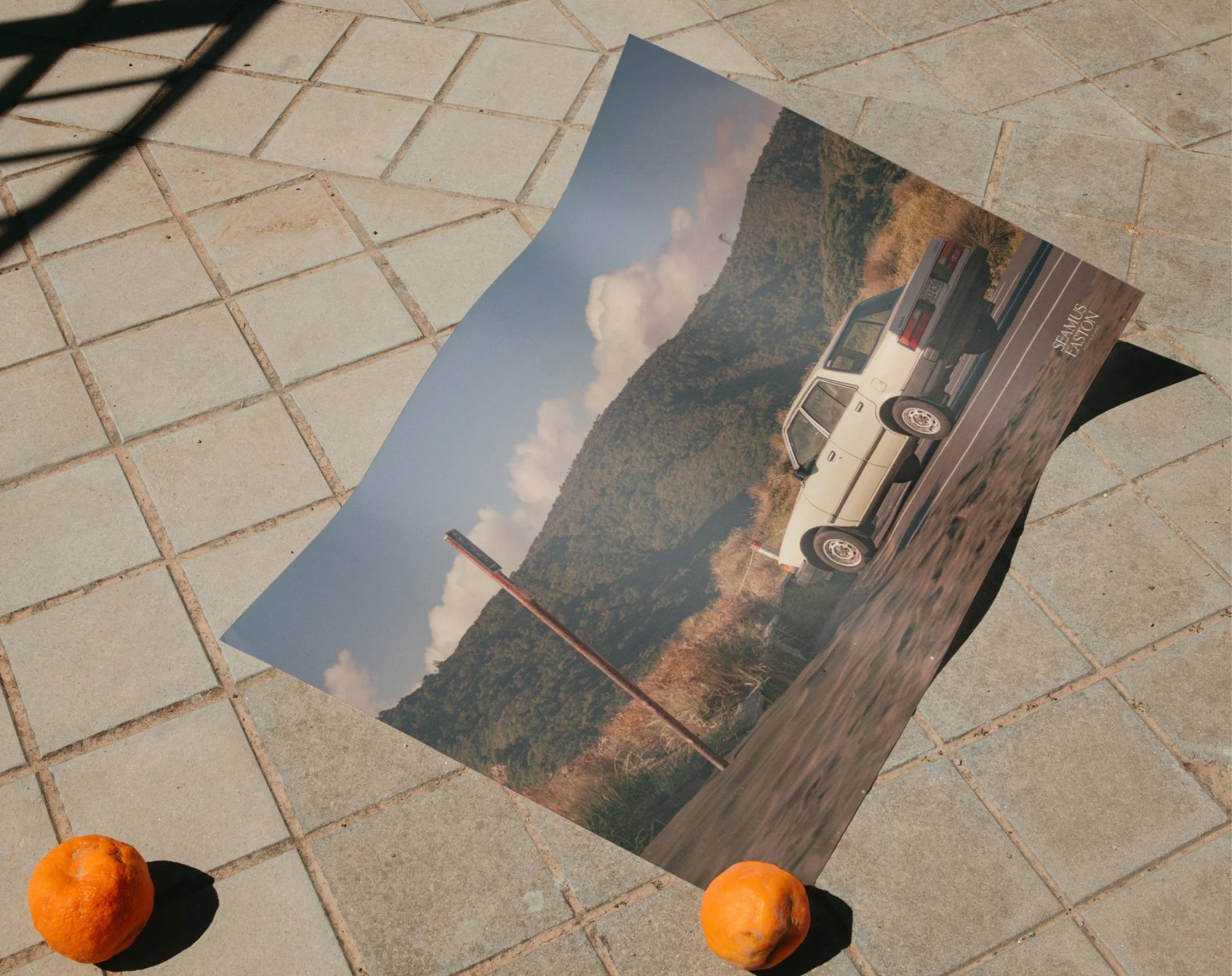 A poster of a vintage Se passage car in a mountainous landscape on a tiled patio, with two oranges on either side.