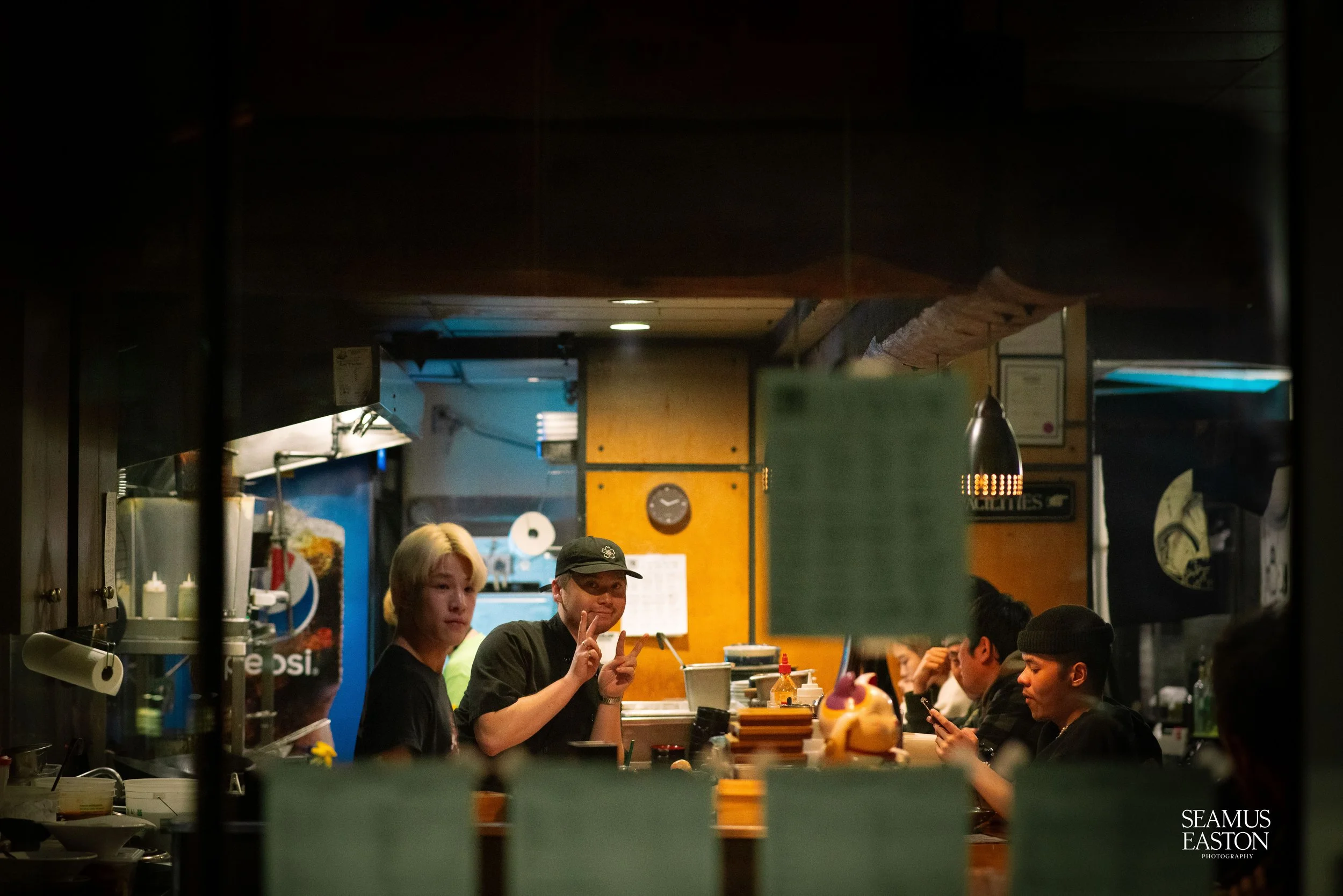 Inside a busy restaurant kitchen with staff and customers, view through a serving window, two staff members near the center, one making a peace sign and the other looking at the camera, various kitchen items and framed certificates on the wall.