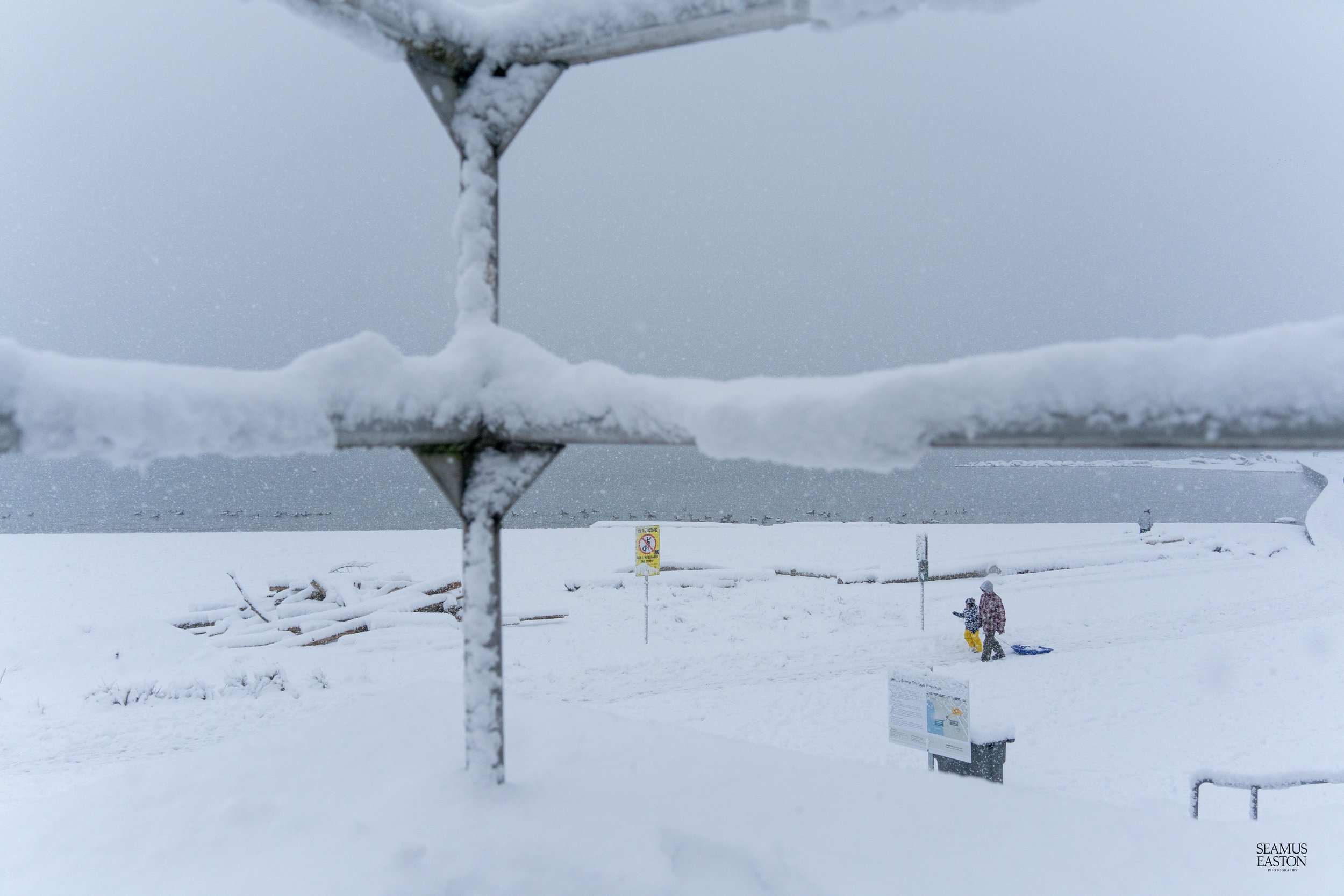 Snow-covered outdoor scene with two people, one adult and one child, near a sled, in a snowstorm. A sign and an informational board are visible in the background.