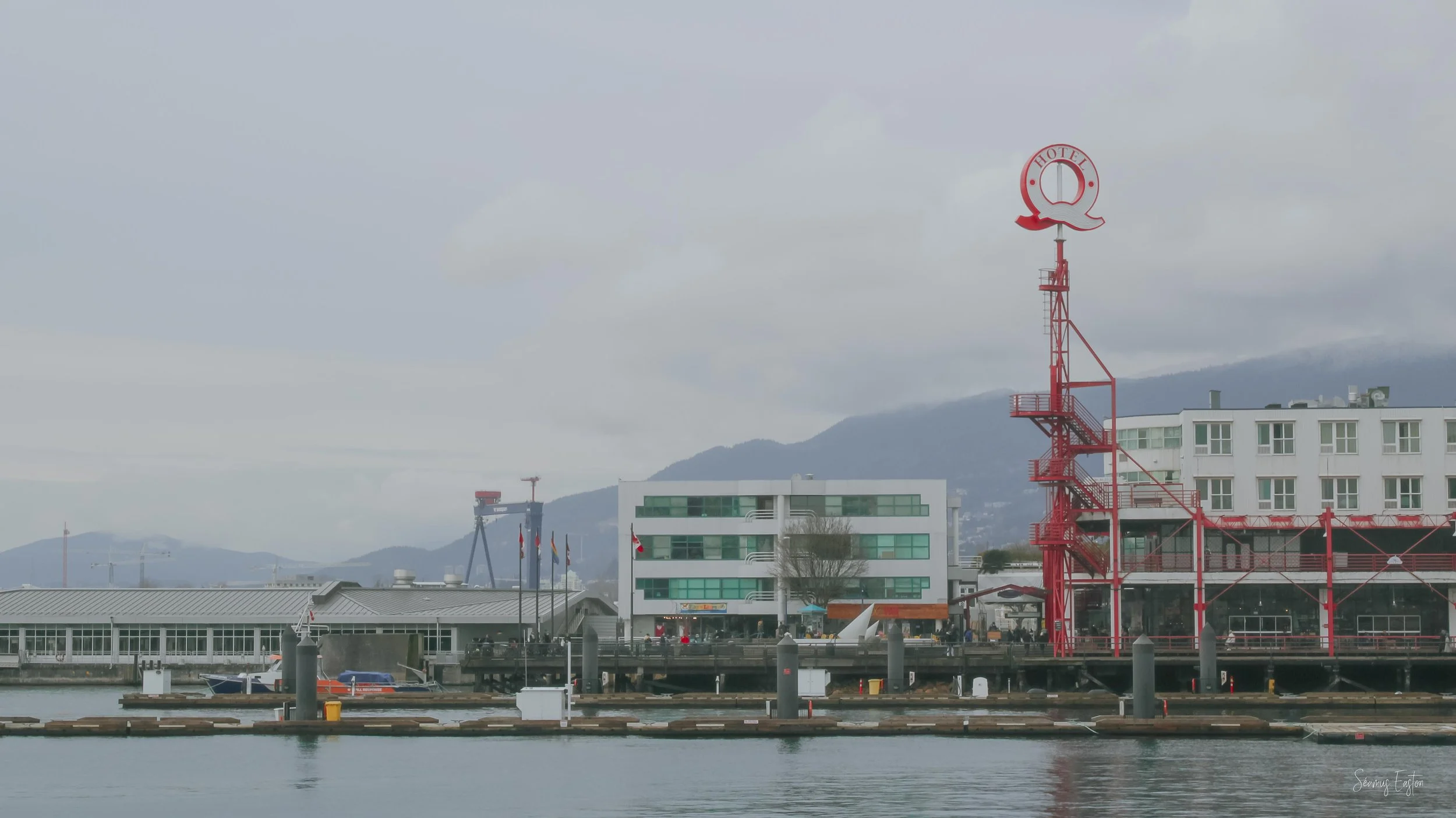 View of a harbor with boats, modern buildings, and a distinctive red lighthouse with the word 'HOTEL' on top, mountains in the background, and cloudy sky.