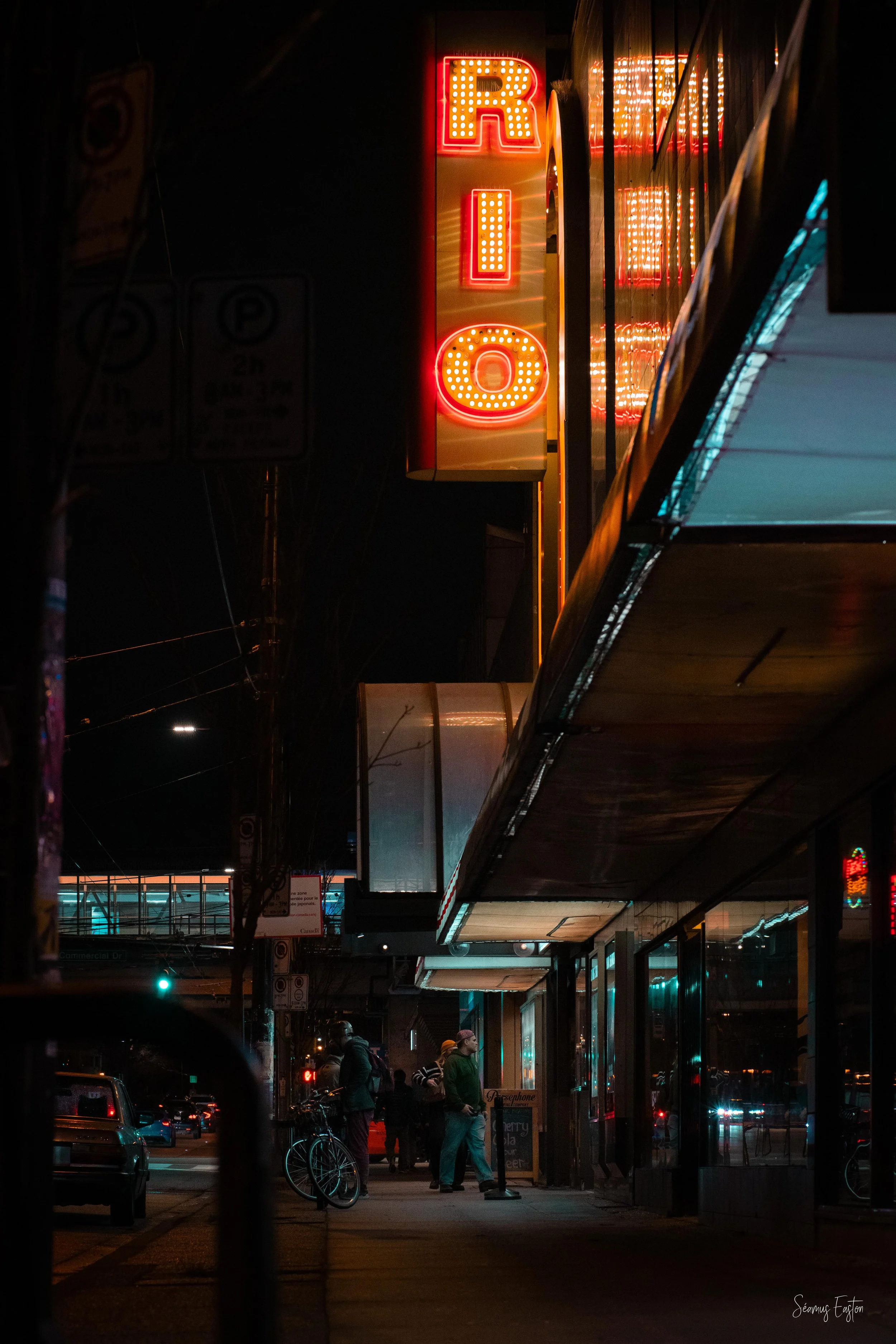Night street scene with a vertical illuminated sign reading 'RION', people standing outside a storefront, bicycles, and cars parked along the street.