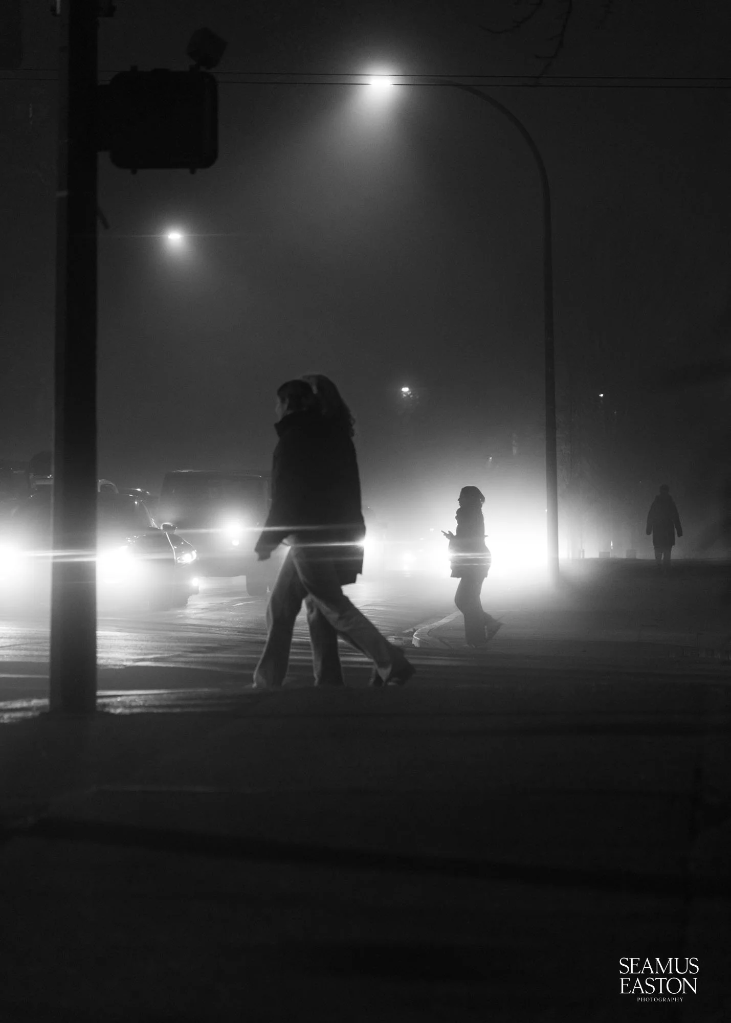 People crossing a foggy street at night with headlights shining behind them, black and white photography.
