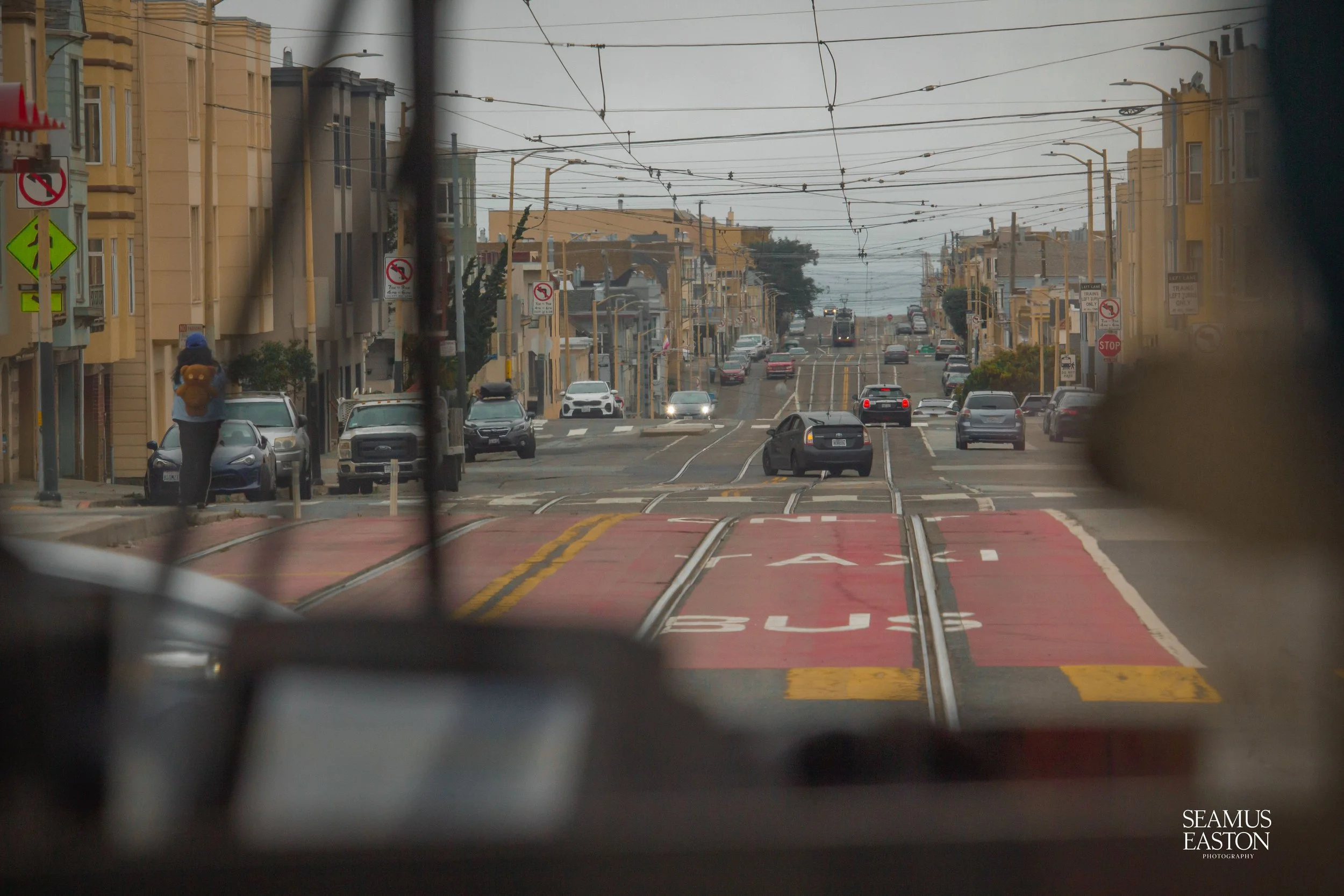 City street viewed from inside a vehicle with tram tracks, cars, buildings, street signs, and an overcast sky.